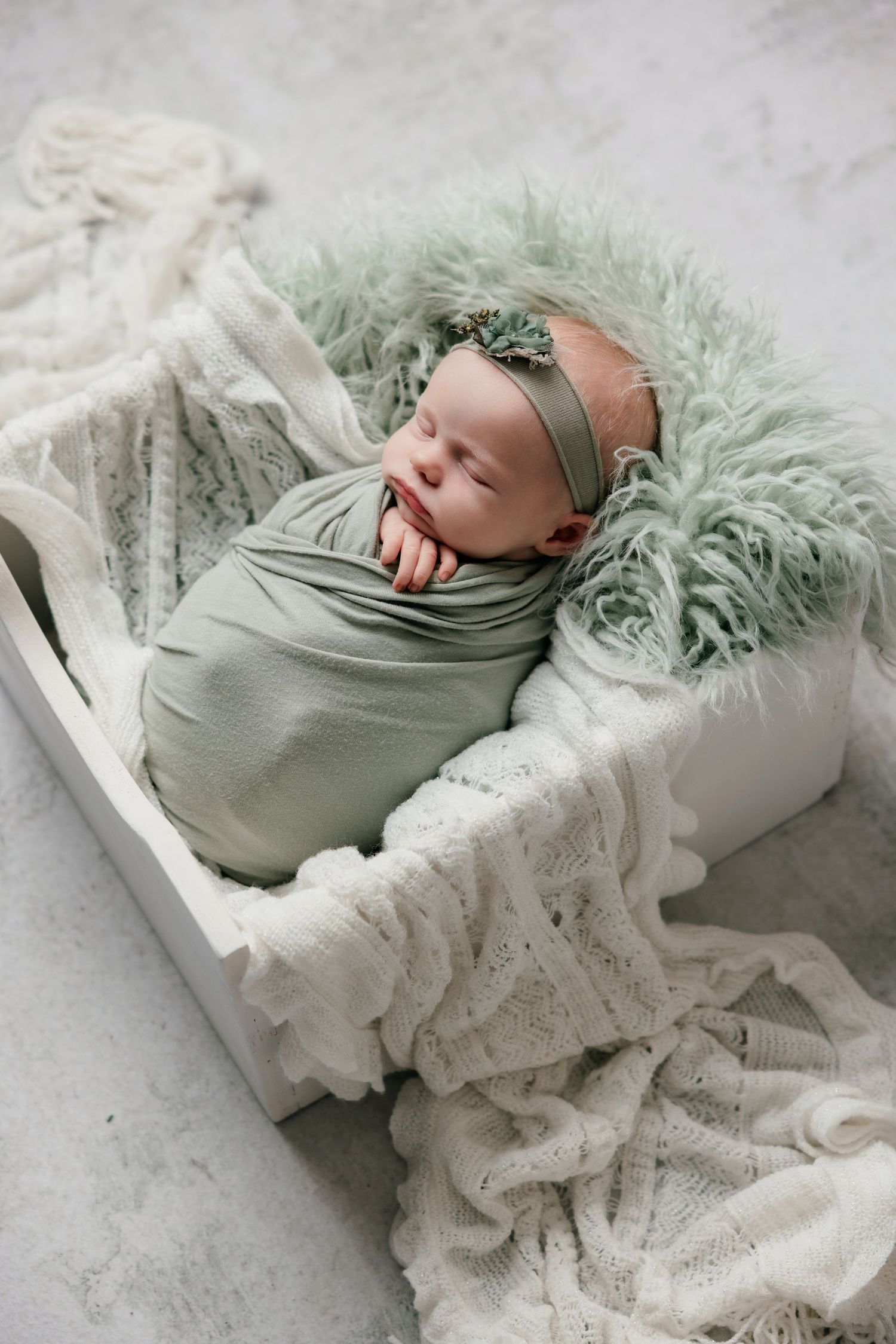 Newborn baby swaddled in sage green wrap with floral headband sleeping peacefully in a white wooden box.