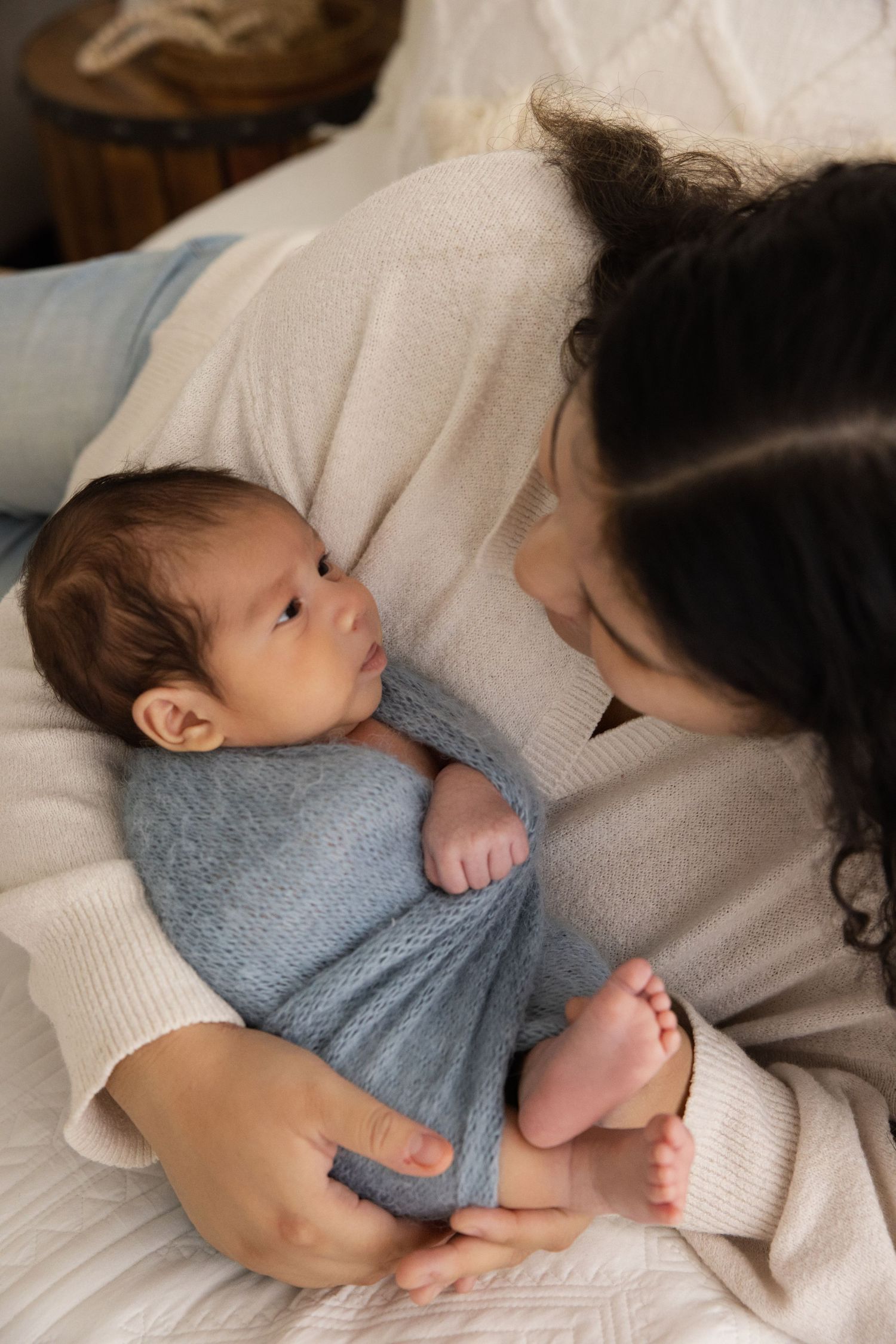 A mother tenderly gazes at her newborn baby wrapped in a blue knit blanket while lying on a white bed.