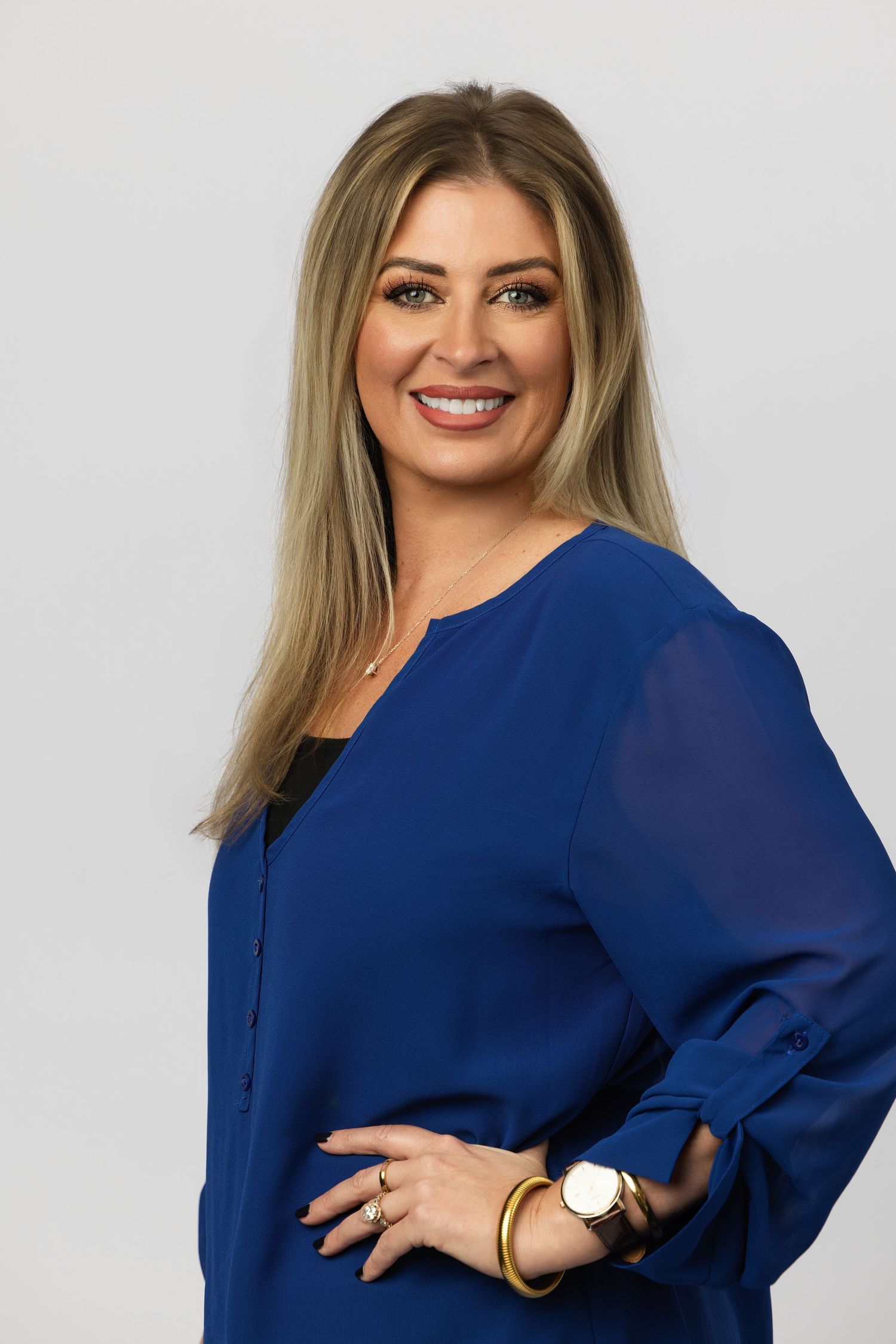 Professional woman in blue blouse smiling confidently, wearing gold accessories, against a light gray background.