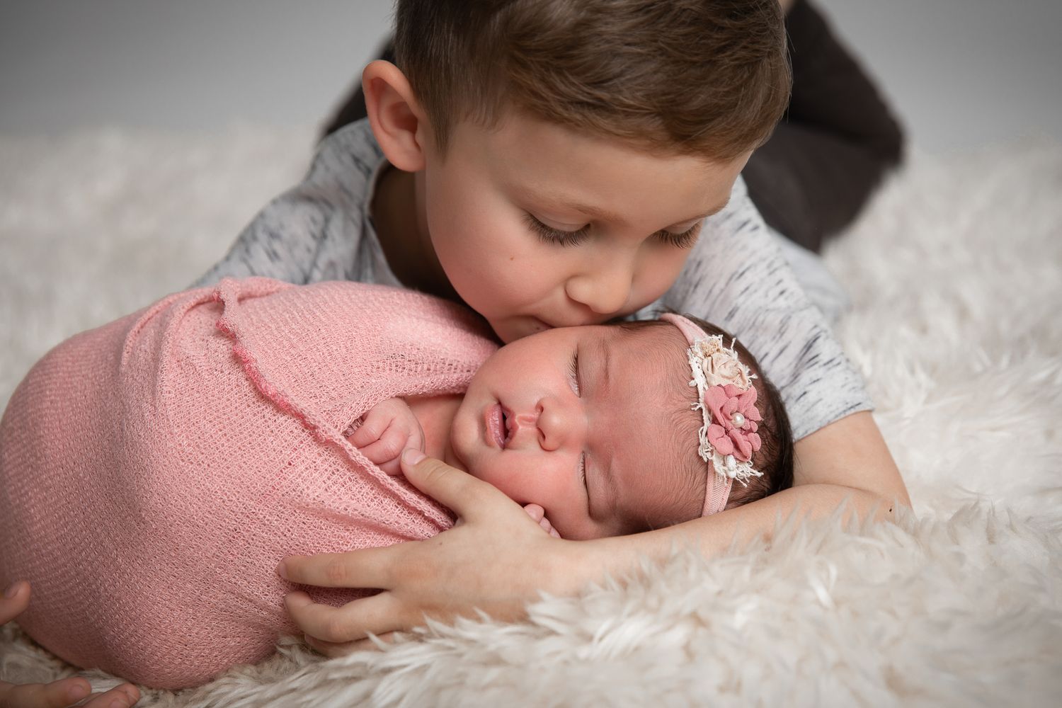 Young boy gently kissing sleeping newborn baby sister wrapped in pink on a soft white fur blanket.