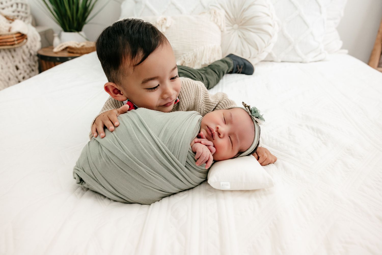 A toddler boy smiles while hugging a swaddled newborn baby sibling lying on a white bed.