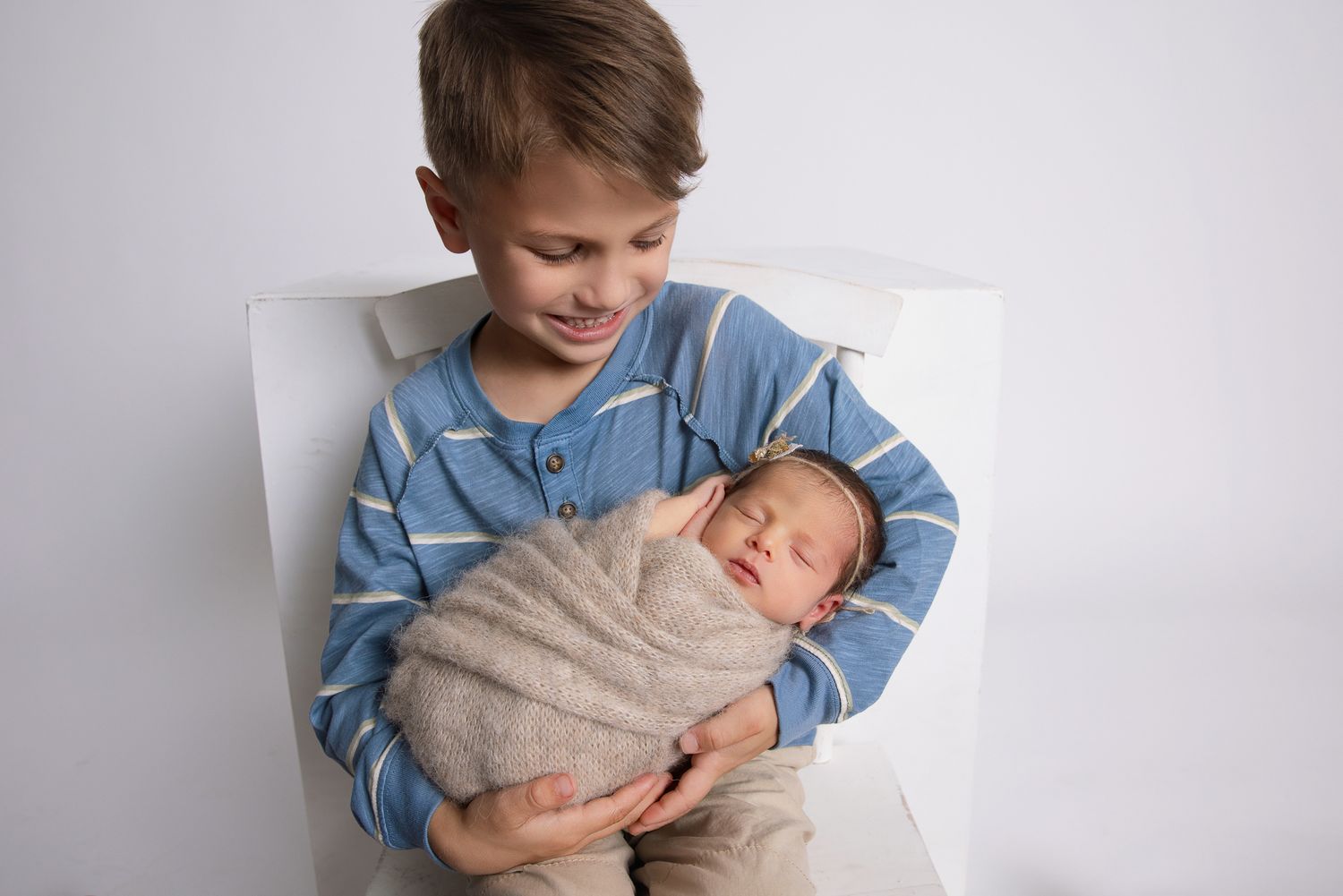 Older boy in blue striped shirt smiling while gently holding a sleeping newborn baby wrapped in a beige swaddle.