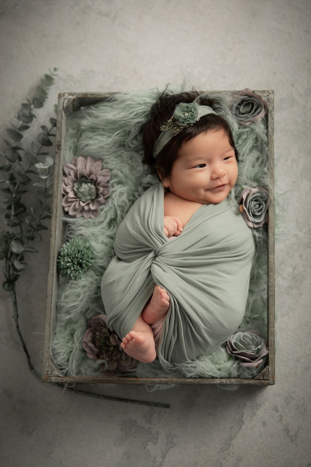 Smiling newborn baby wrapped in sage green swaddle, lying in a wooden box with floral decorations and green fur.