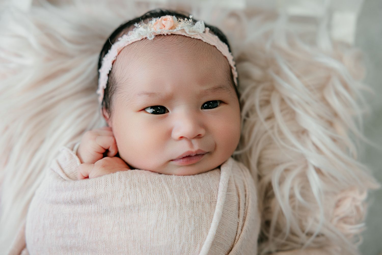 Newborn baby girl wrapped in pink swaddle with floral headband lying on fluffy white fur backdrop.
