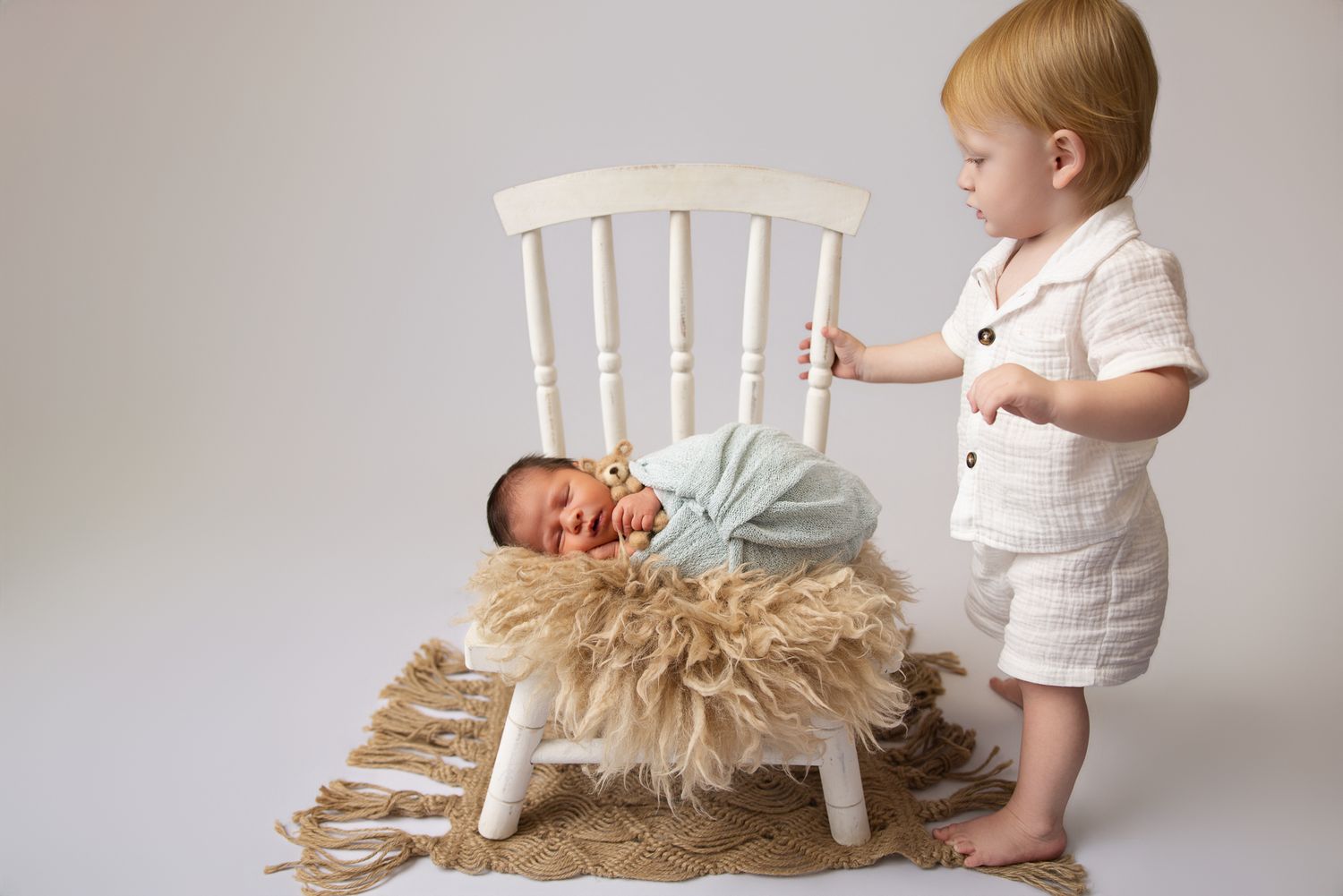 Toddler boy in linen outfit stands beside a white chair holding a sleeping newborn baby wrapped in mint on a fur rug.