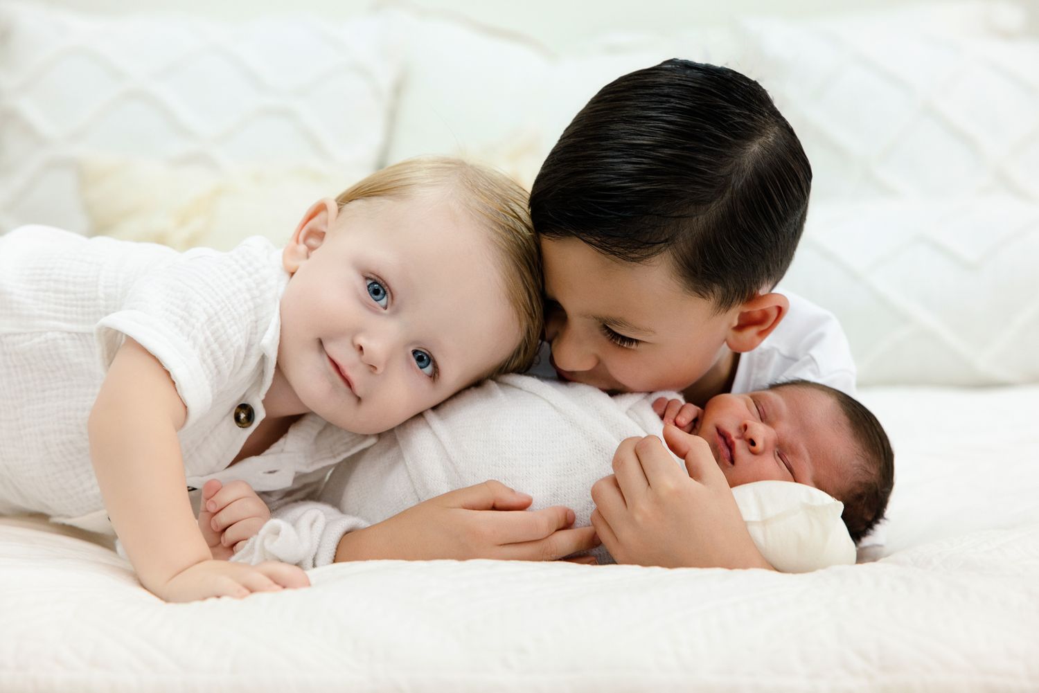 Two young siblings lovingly cuddle a newborn baby on a white bed, wearing matching white outfits.
