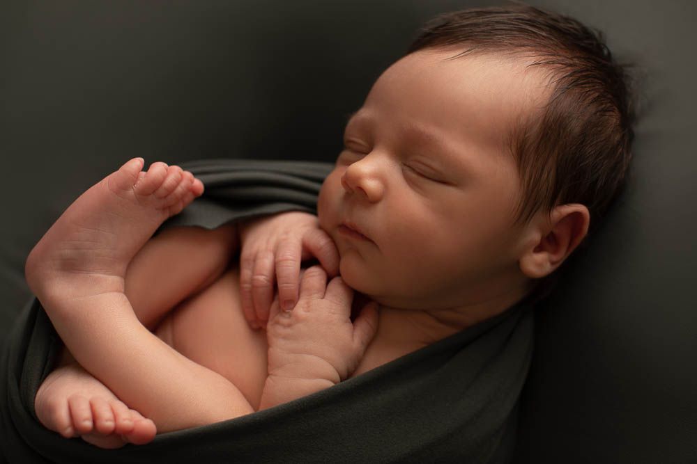 Peaceful newborn baby sleeping wrapped in a dark green cloth, curled up in a serene studio portrait.