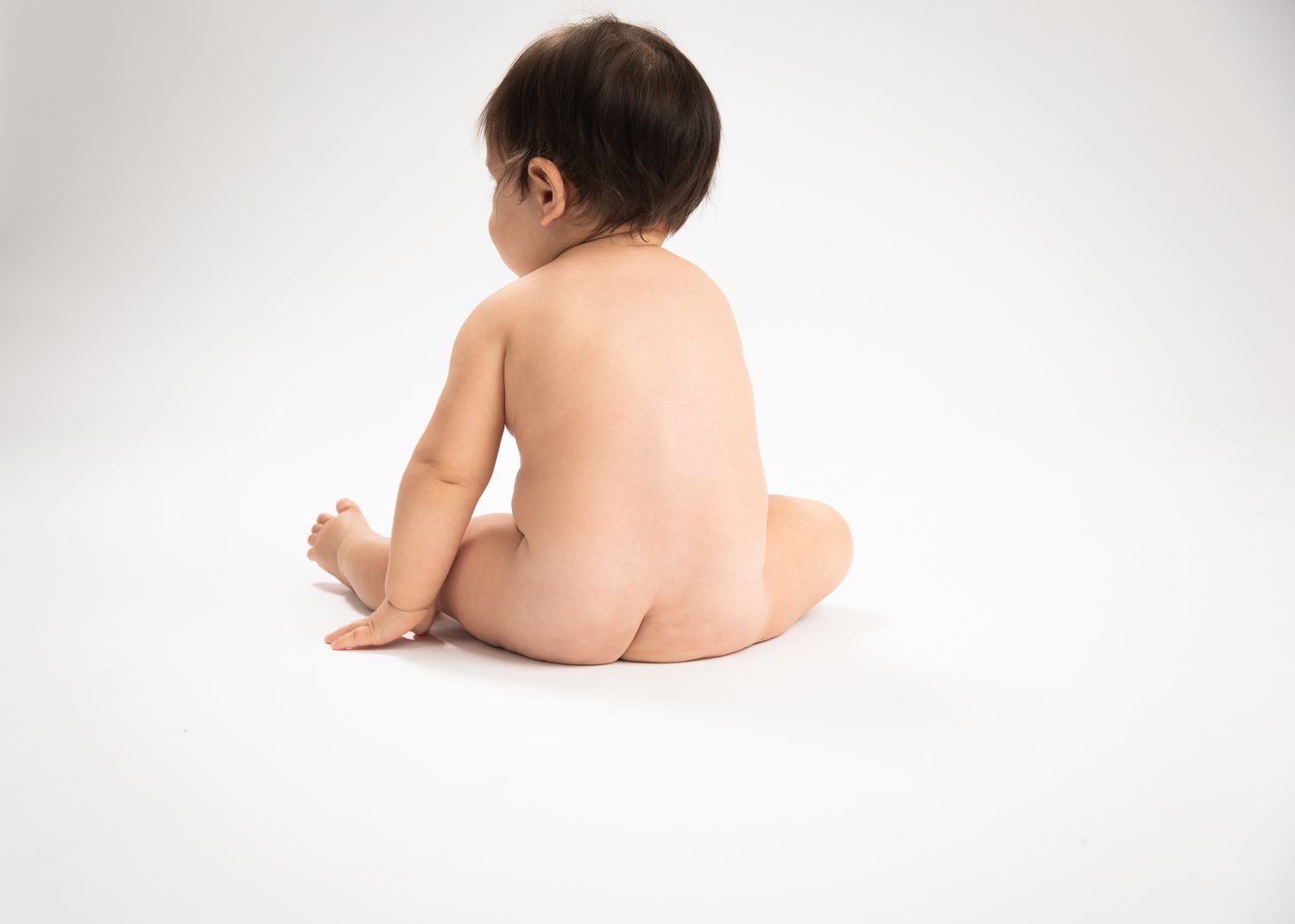 Naked baby sitting with back to camera on white background in studio portrait.