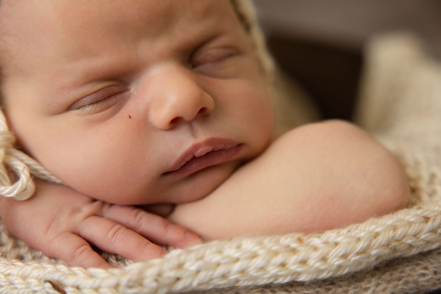 Peaceful newborn baby sleeping with tiny hands tucked under chin, wrapped in a soft cream knit blanket.