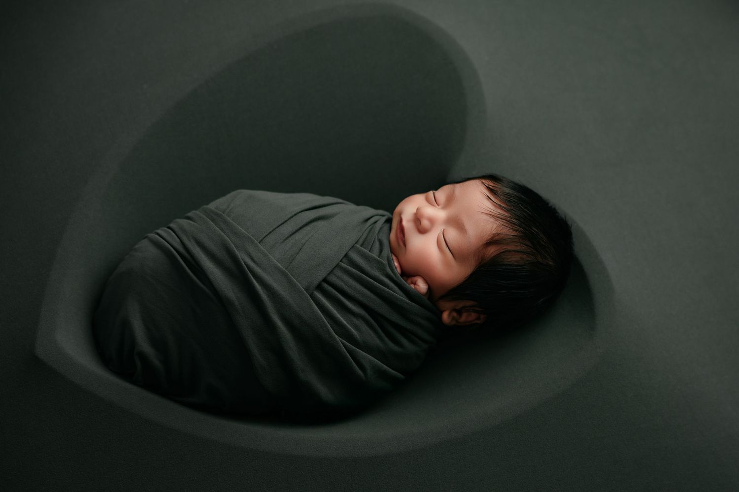 Newborn baby swaddled in dark green wrap sleeping peacefully inside a heart-shaped bowl prop.