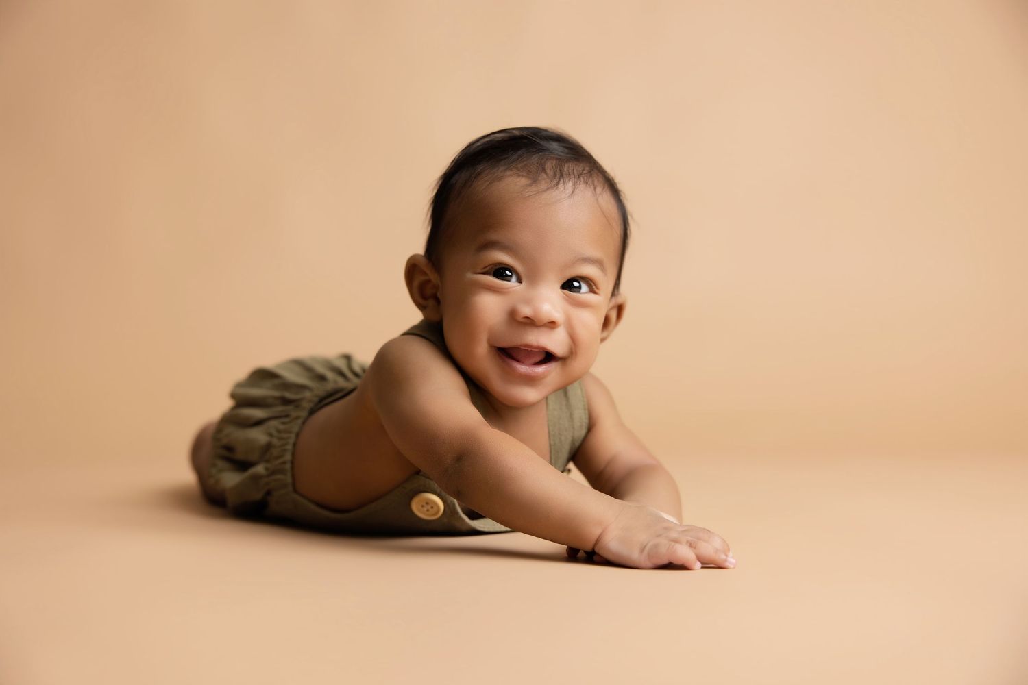Smiling baby in olive green romper doing tummy time on beige background.