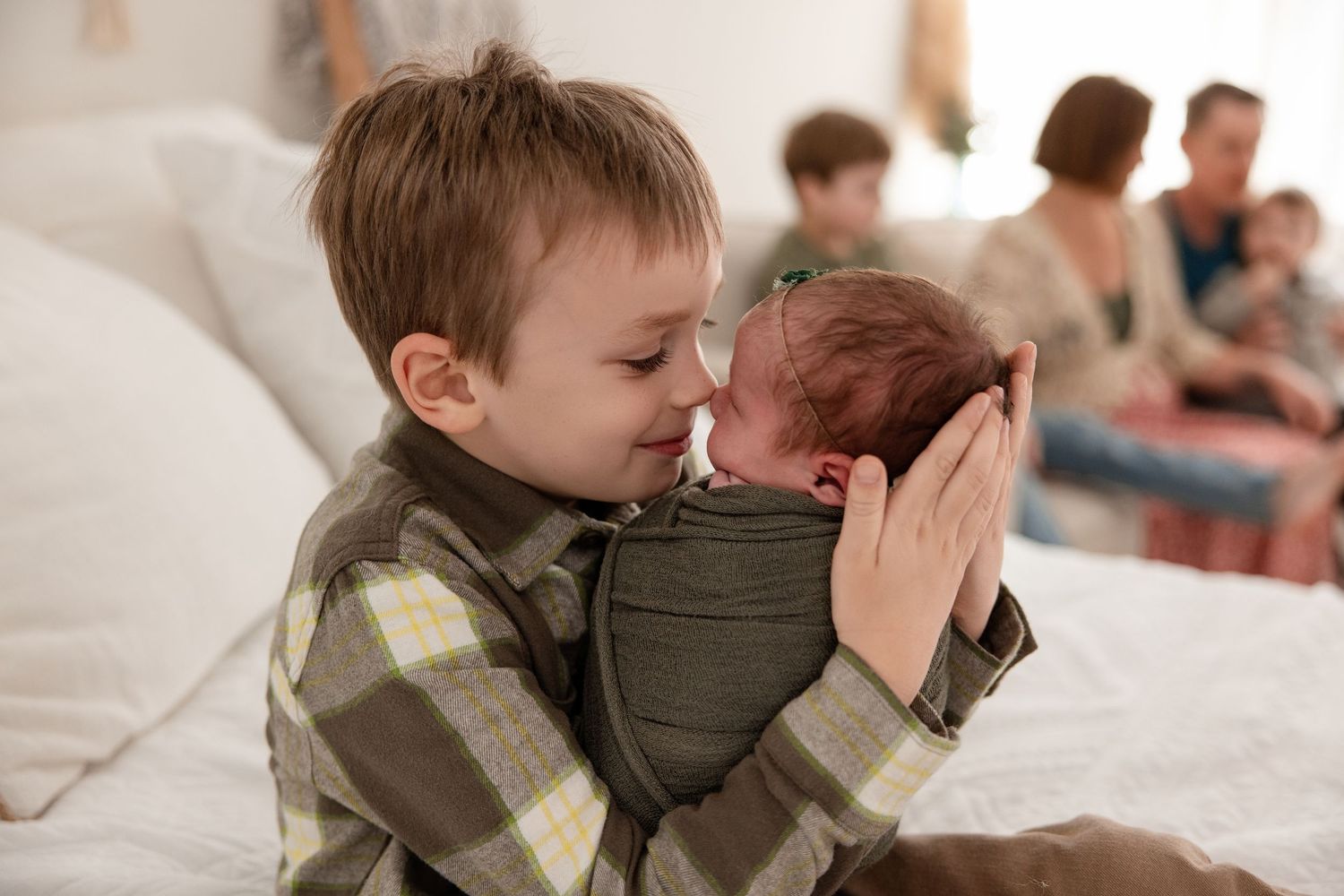 Young boy tenderly holds newborn baby while family sits in background on white bed.