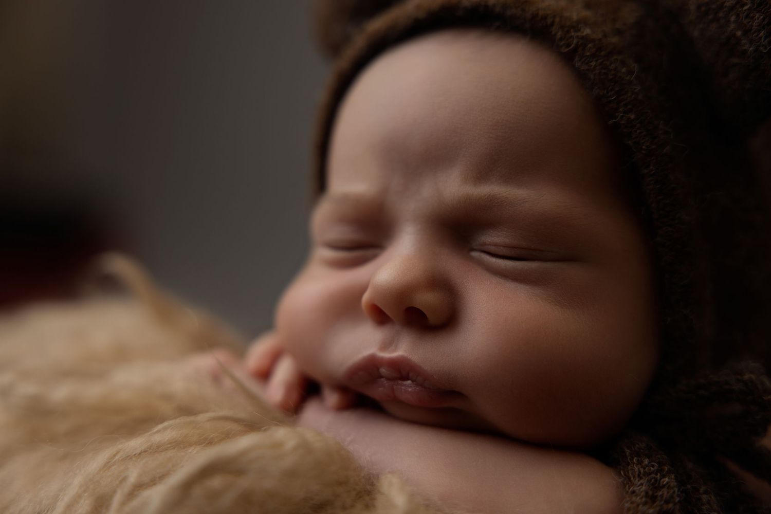 Peaceful newborn baby sleeping soundly, wearing a cozy brown knit bonnet, with tiny hands tucked under chin.