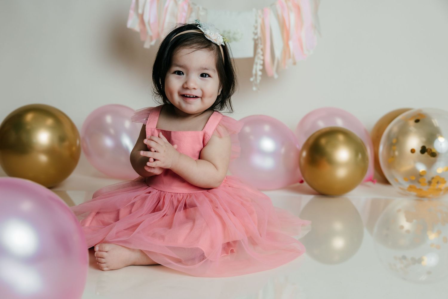 Smiling baby girl in pink tutu dress sitting on floor surrounded by pink and gold balloons at birthday party.