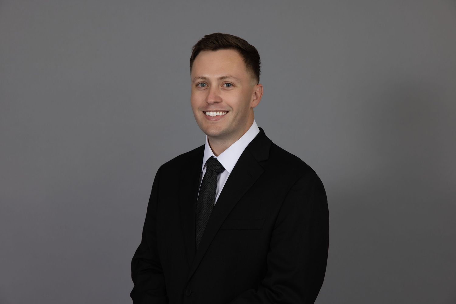 Professional man in black suit and tie smiling confidently against a gray studio background.