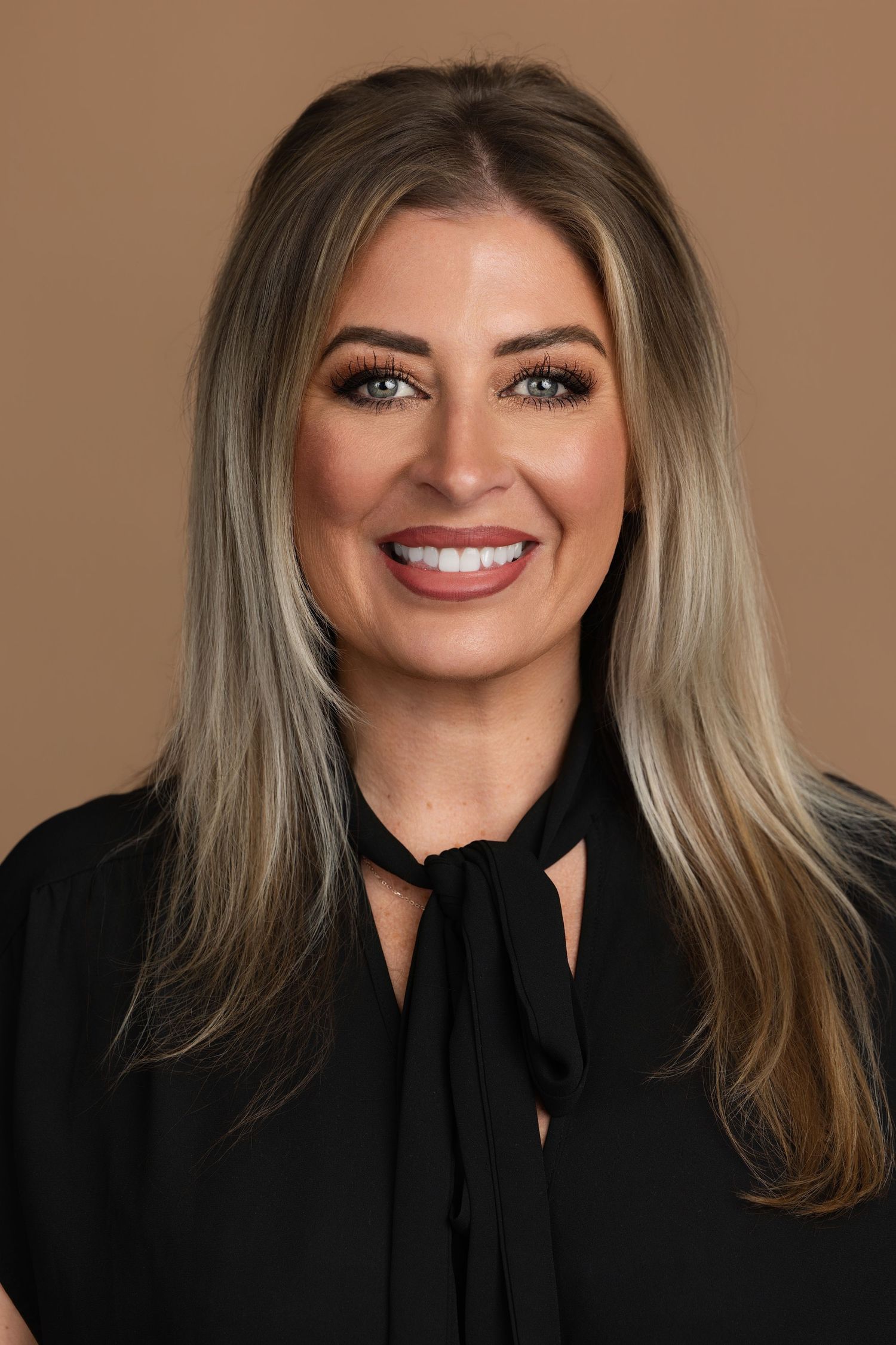 Professional headshot of a smiling blonde woman with blue eyes wearing a black blouse against a tan background.