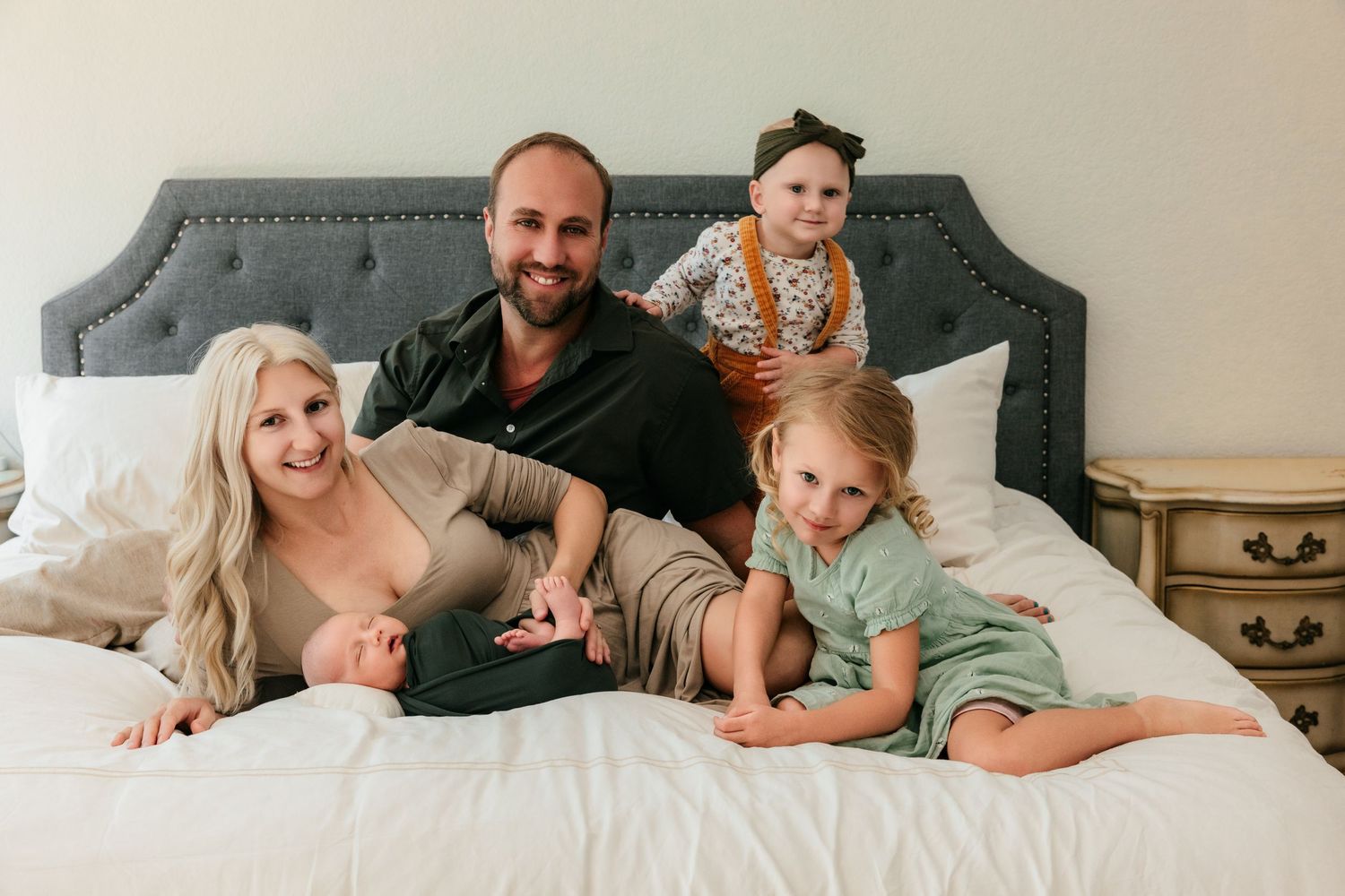 A happy family of four poses together on a white bed, with two young girls and their smiling parents.