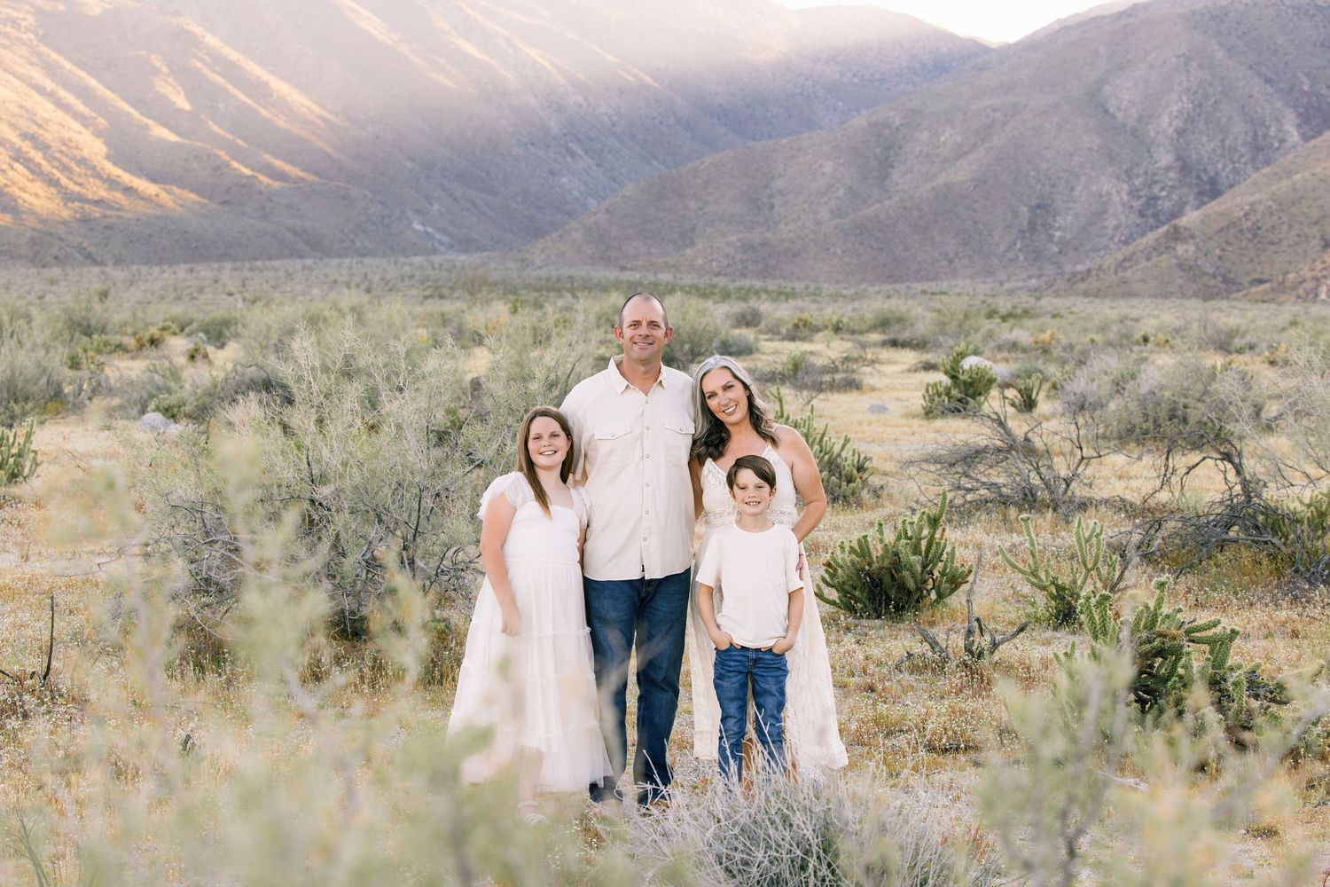 Family of four smiling in desert landscape with mountain backdrop during golden hour photography session.