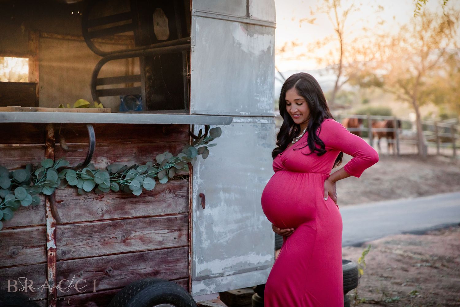 Pregnant woman in a pink maternity dress holding her baby bump beside a rustic wooden trailer outdoors.
