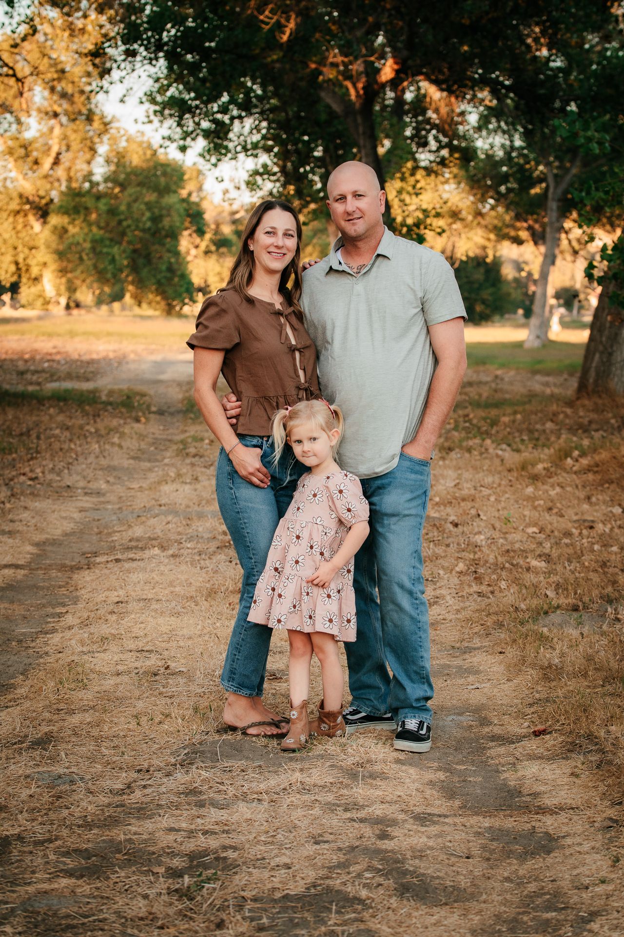 A couple and their young blonde daughter pose on a dirt path surrounded by golden trees at sunset.