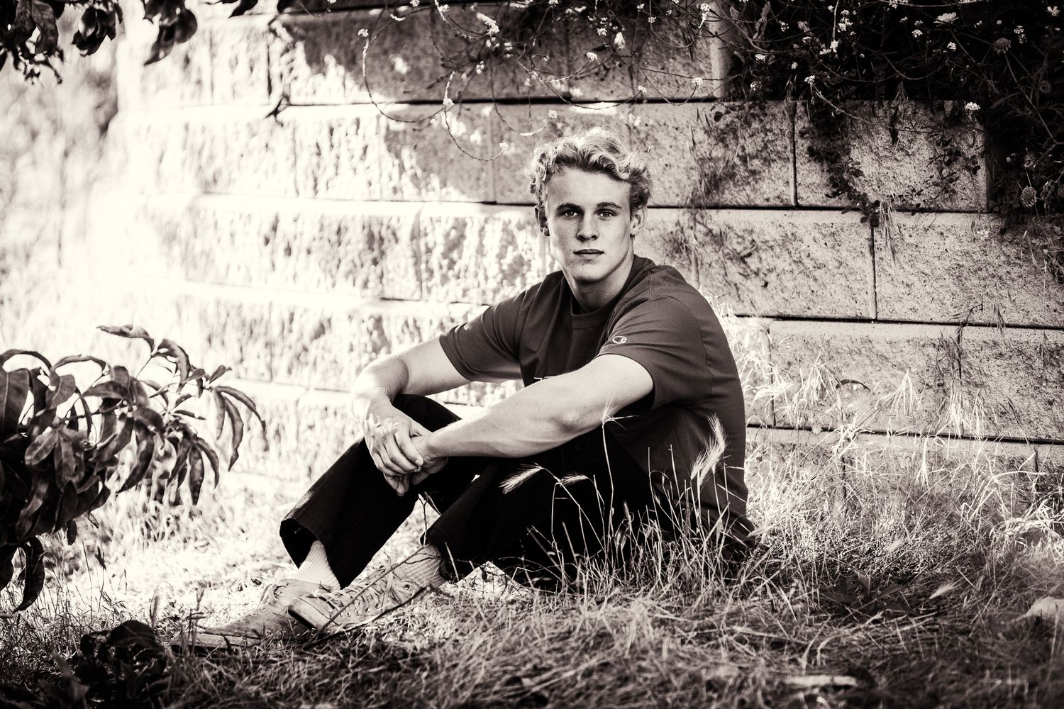 Black and white portrait of young blond man sitting against stone wall in grass.