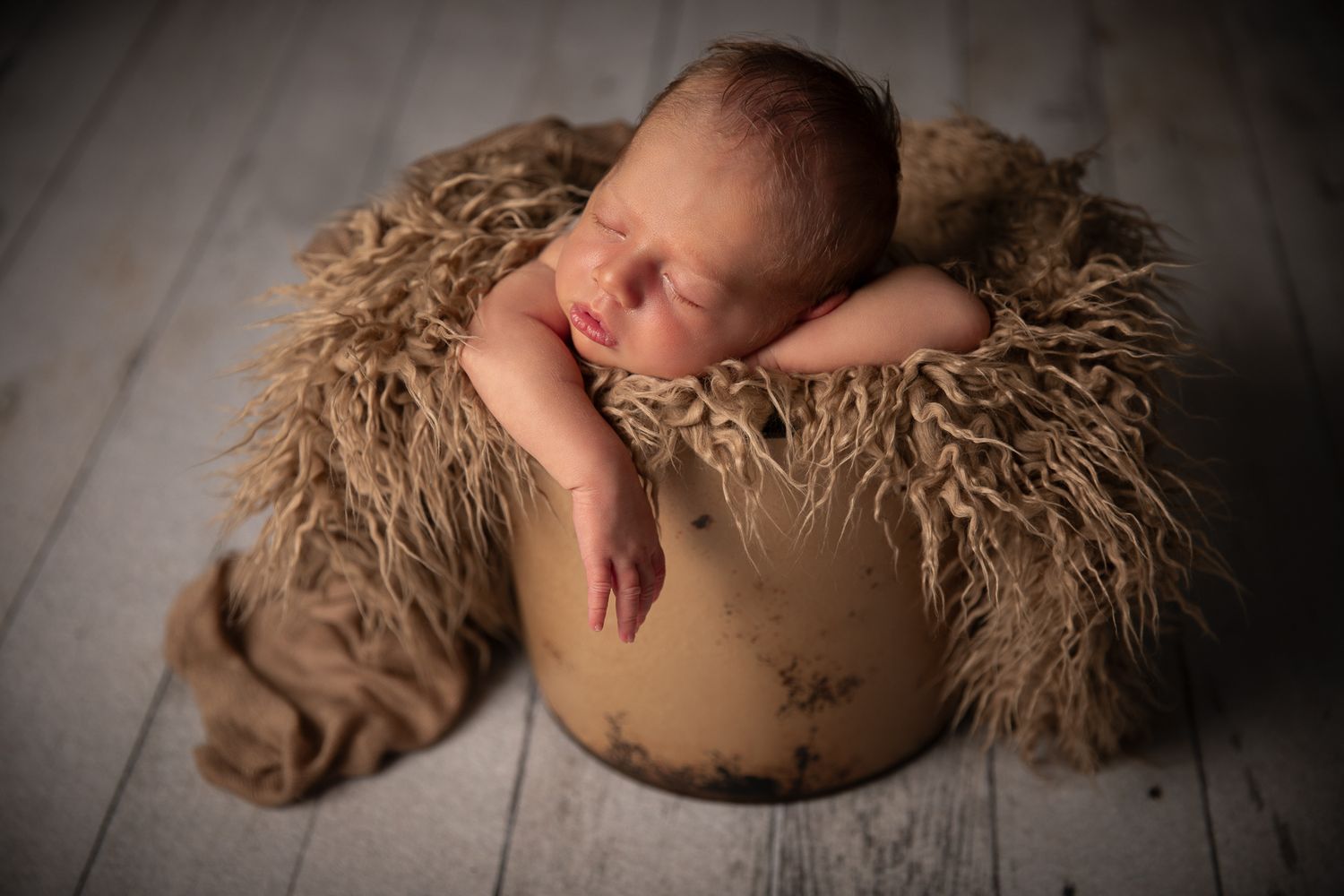 Sleeping newborn baby nestled in a rustic bucket with soft brown fur, studio portrait.