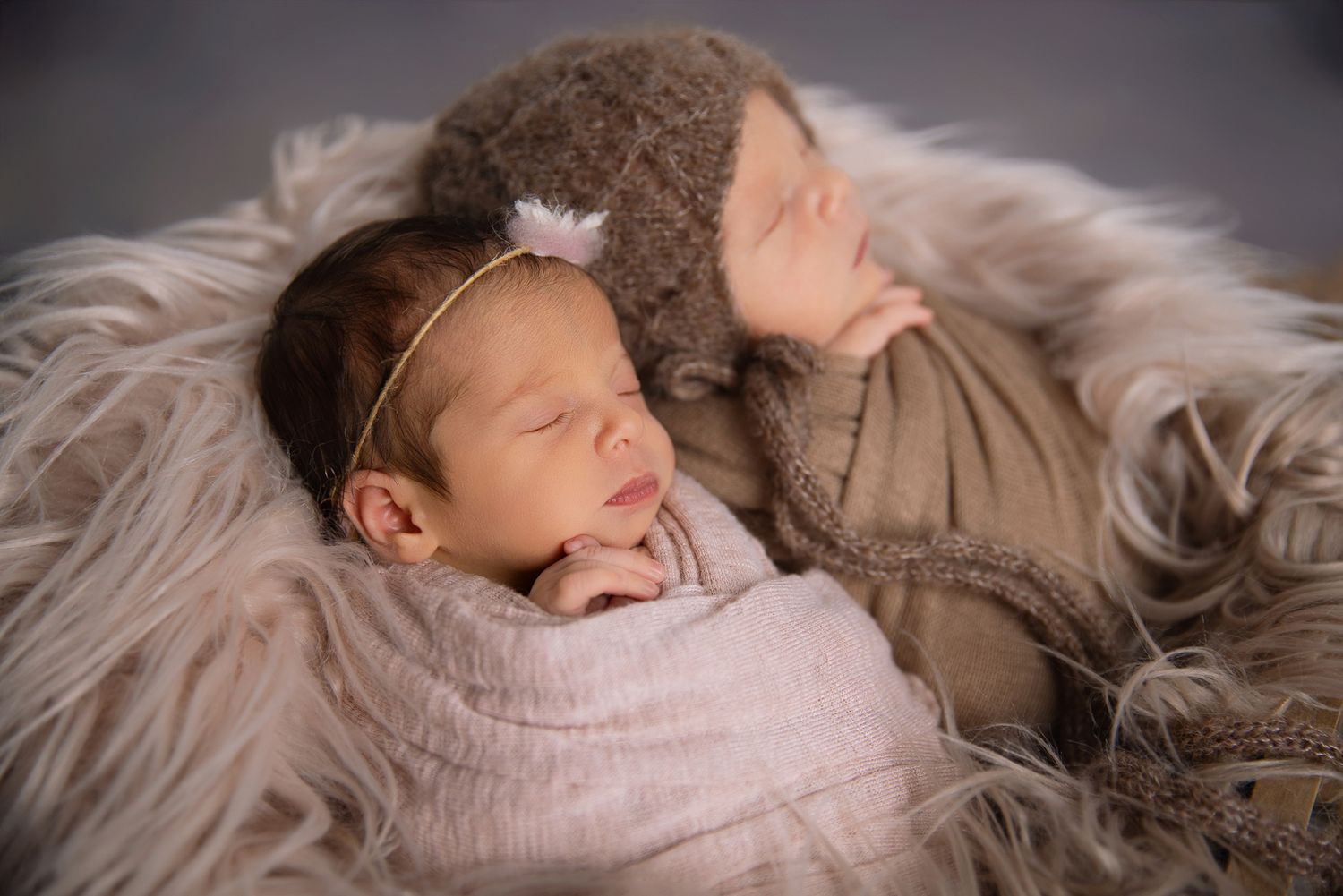 Twin newborns sleeping together on fluffy fur, one in pink wrap with gold headband, one in brown hat. Fallbrook Newborn Photographer