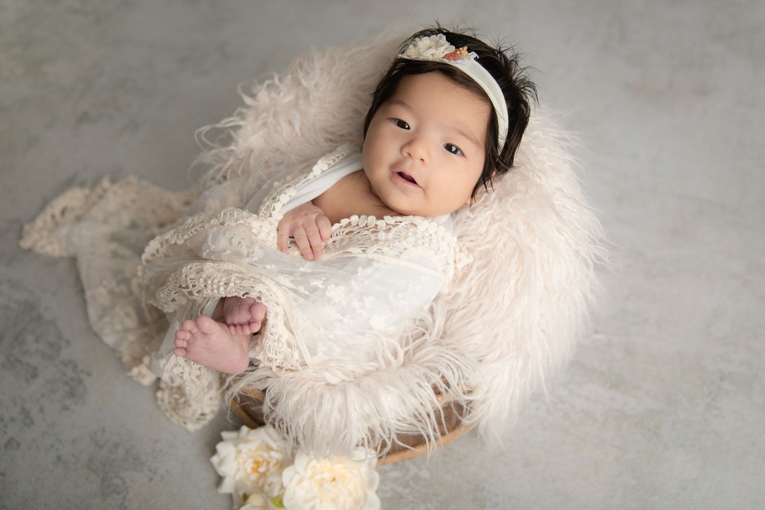 Newborn baby girl in white lace romper resting in fluffy cream basket with floral headband for studio portrait.