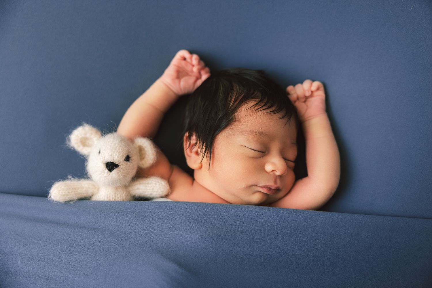 Peaceful newborn baby sleeping on blue blanket with white stuffed bunny toy, arms raised above head.