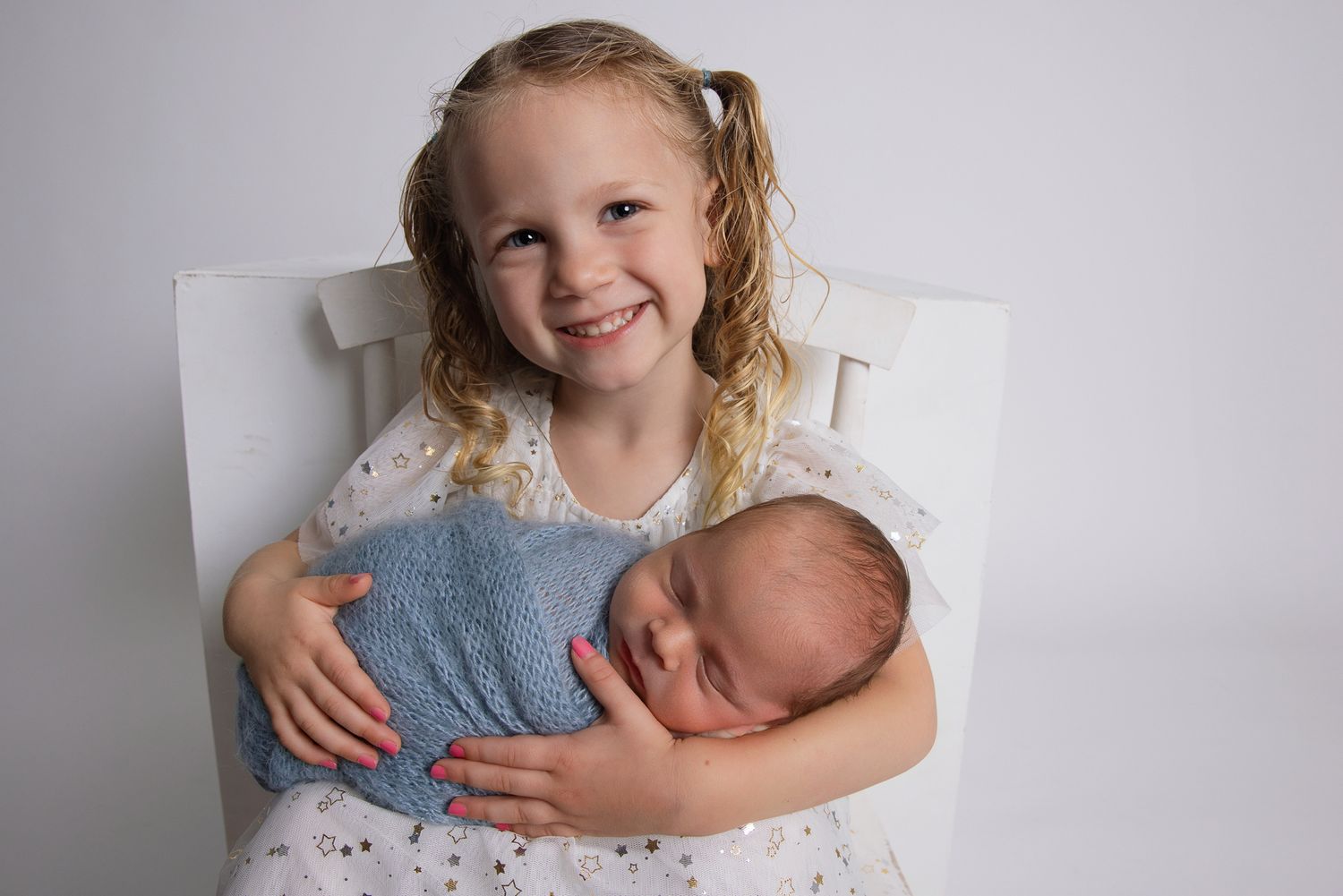 Smiling young girl with blonde pigtails holds a sleeping newborn baby wrapped in a blue swaddle on a white chair.