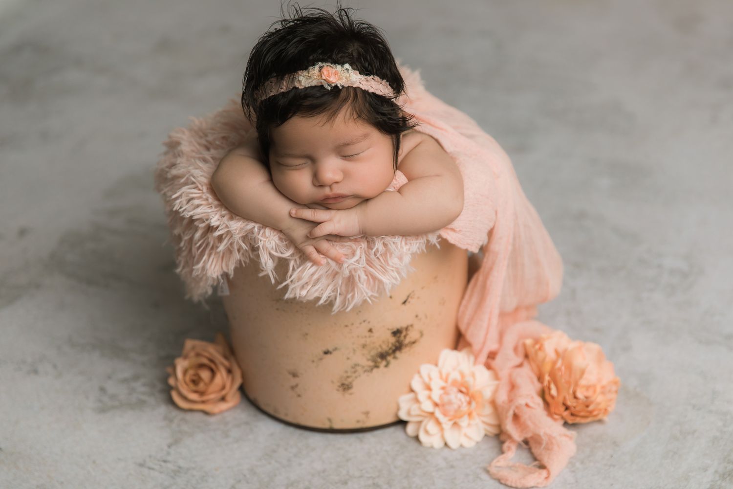 Sleeping newborn baby girl in pink tutu dress and floral headband posed in wooden bucket with peach flowers.