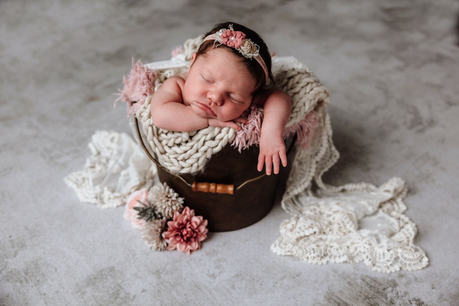 Newborn baby girl sleeping peacefully in a wooden bucket with pink flowers and a floral headband on a gray backdrop.