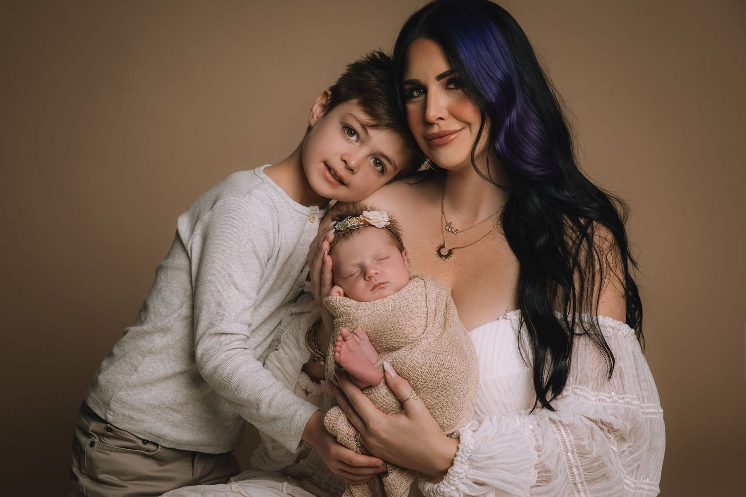 A mother with purple hair holds a swaddled newborn while her young son leans in for a studio family portrait.
