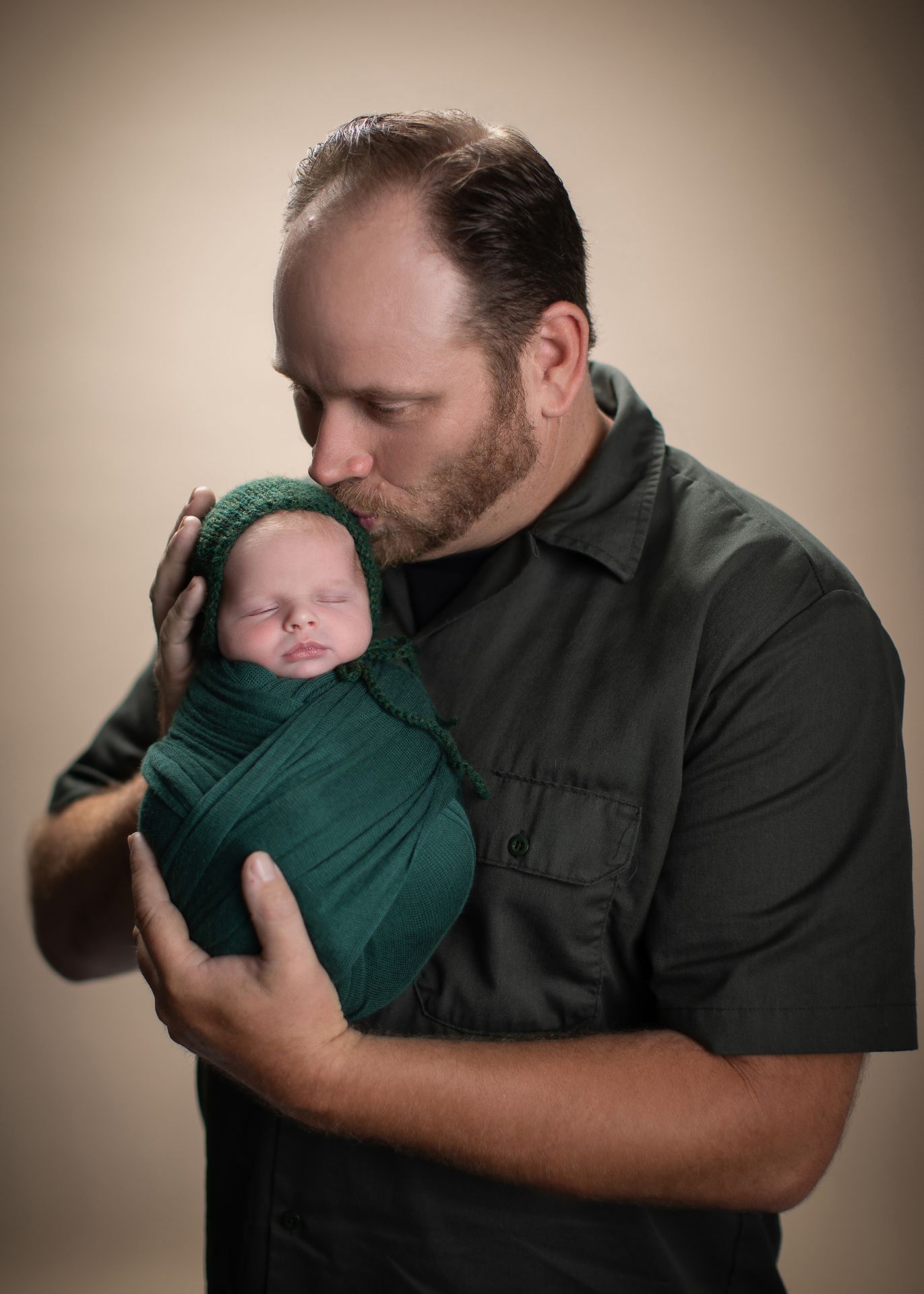 A father tenderly kisses his swaddled newborn baby wrapped in green cloth during a professional portrait session.