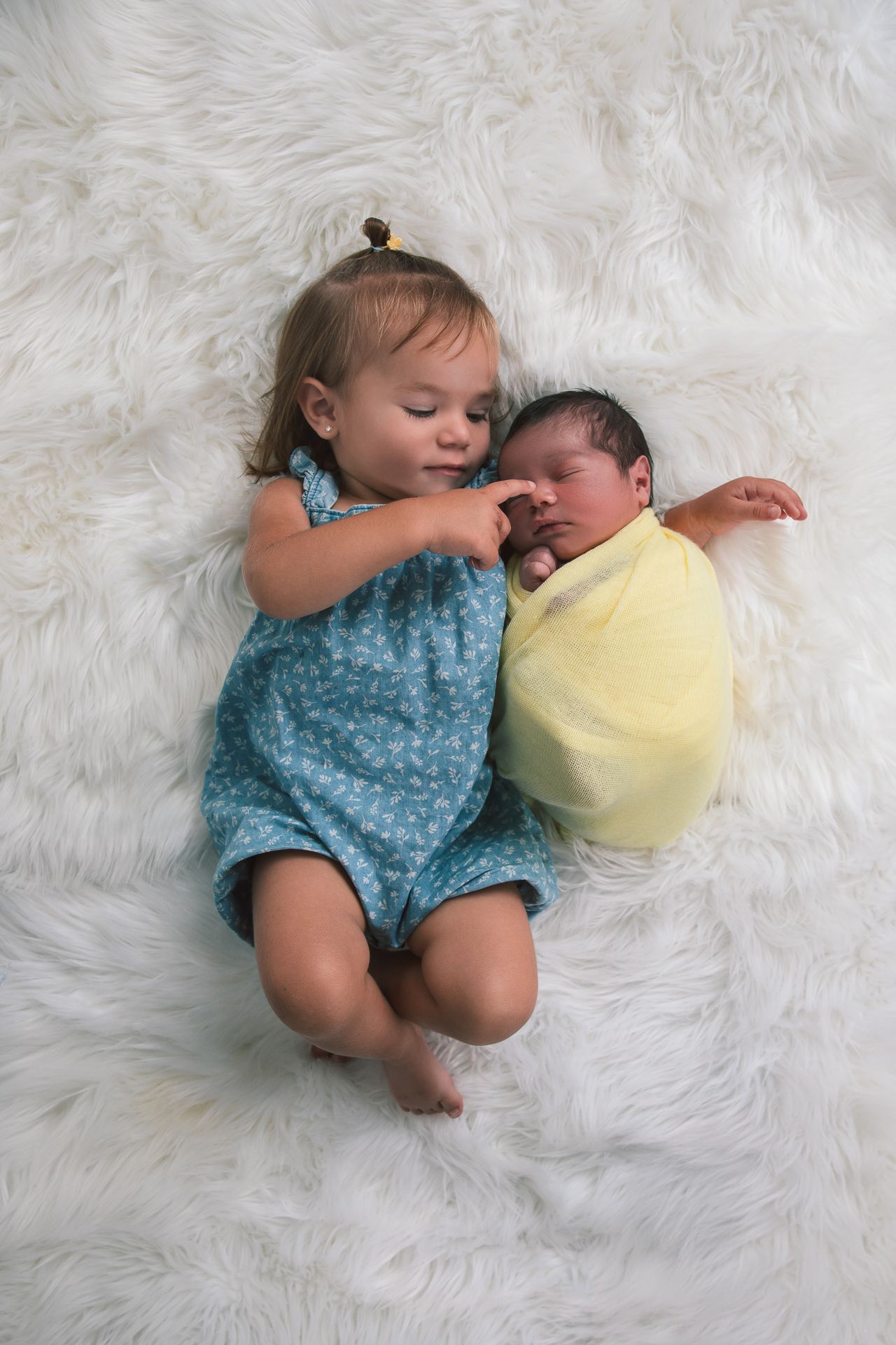 A toddler girl in a blue dress lying next to a swaddled newborn baby on a white fluffy rug.