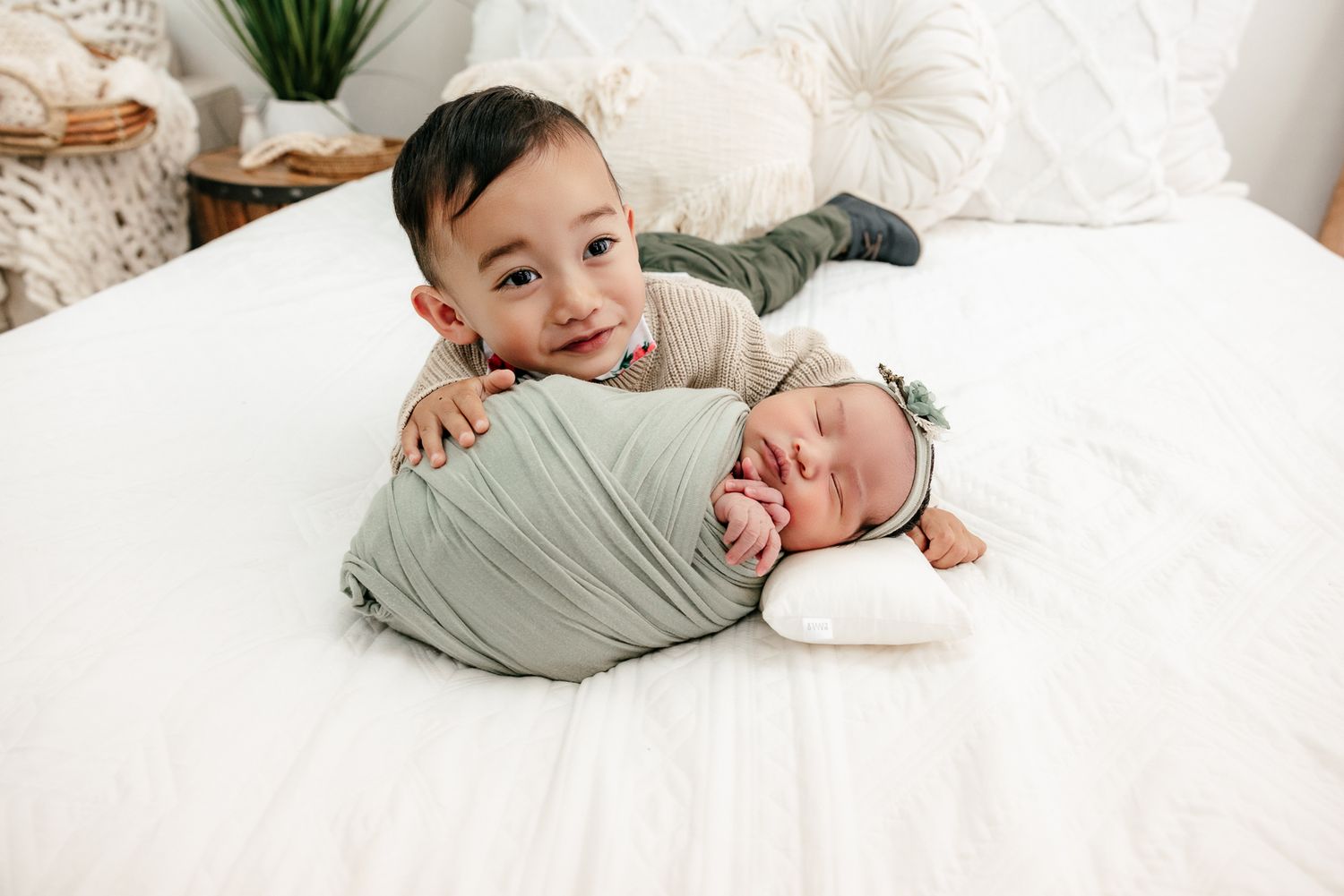 Toddler boy smiling while lying on a white bed next to a swaddled newborn baby in a sage green wrap.