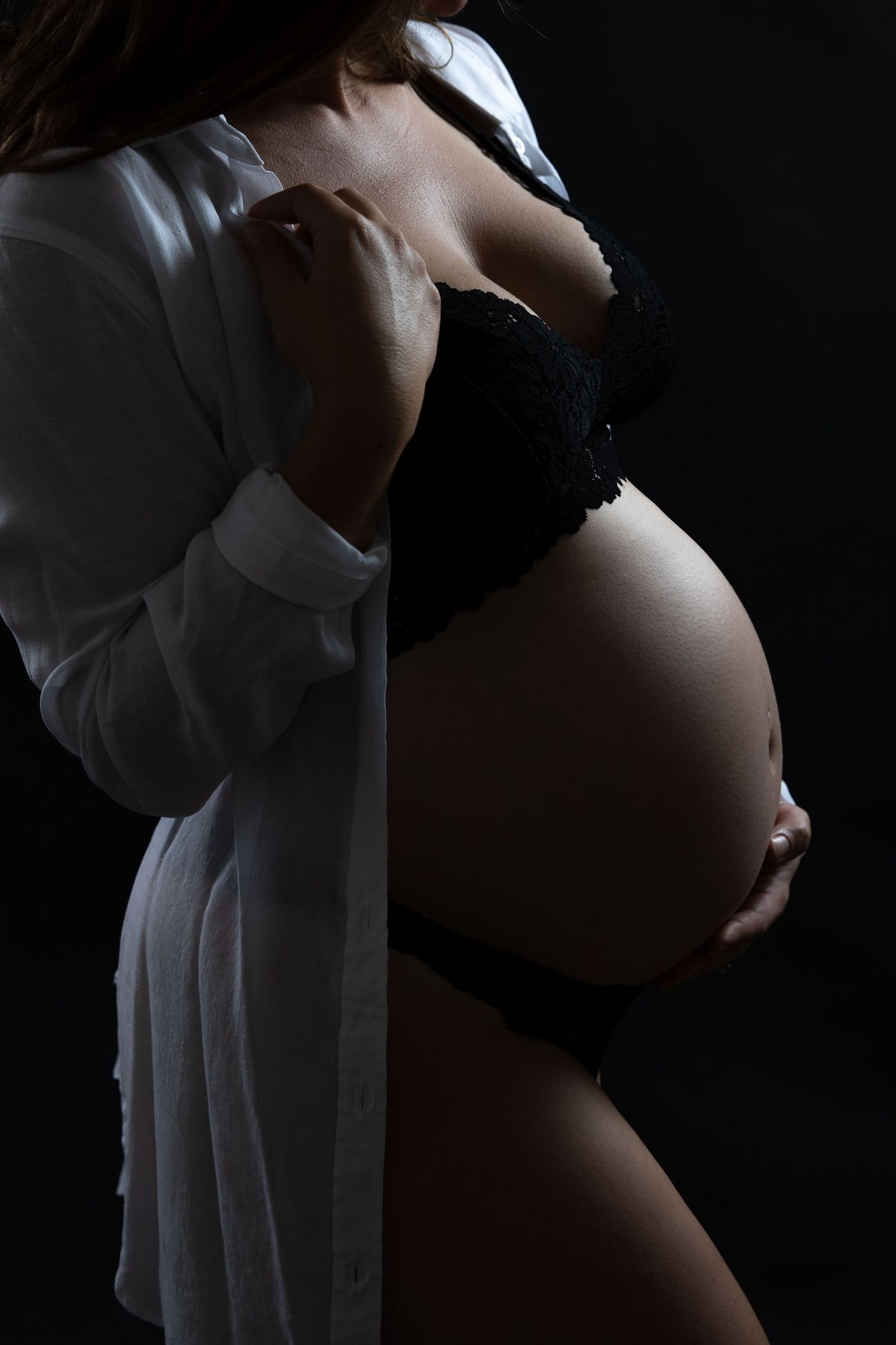 Pregnant woman in black lingerie and white shirt holding baby bump in dark studio portrait.