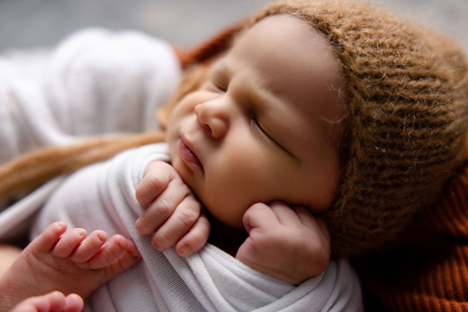 Peaceful newborn baby sleeping in a brown knit hat, wrapped in white swaddle with tiny hands near face.