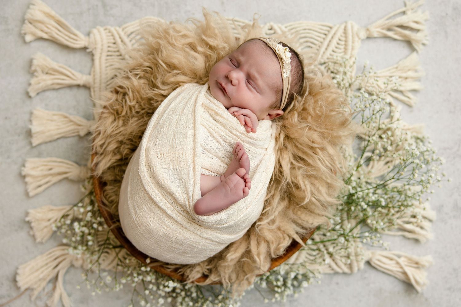Newborn baby swaddled in cream wrap sleeping in a wooden bowl on a fur rug with white flowers.