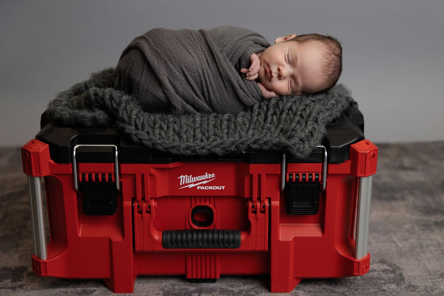 Newborn baby sleeping peacefully in a gray knit nest on top of a red Milwaukee tool case.