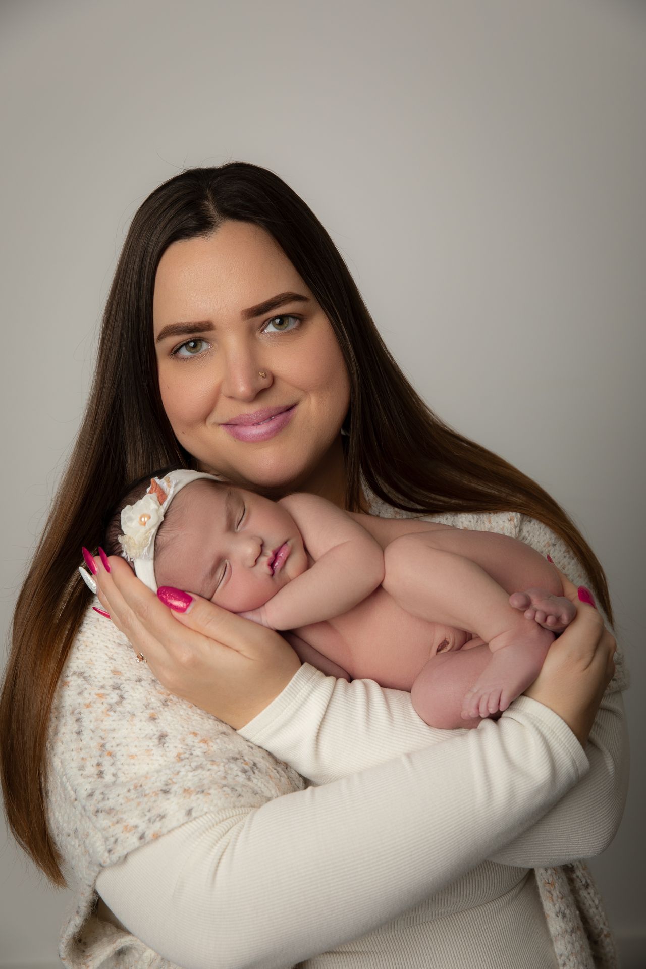 A smiling brunette mother with pink nails gently holds her sleeping newborn baby against her chest in a studio portrait.