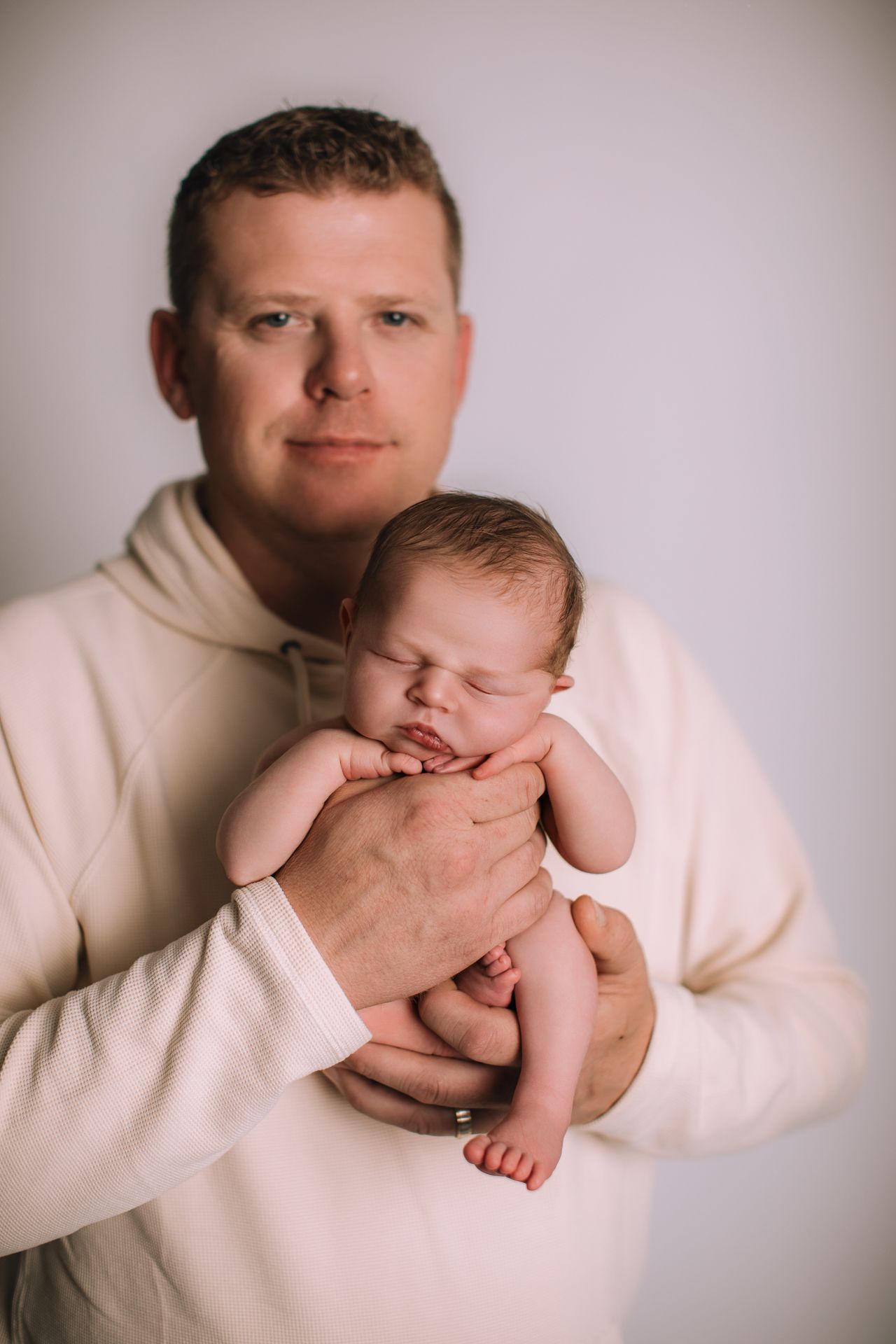 A proud father in a cream hoodie tenderly holds his sleeping newborn baby against a soft white background.