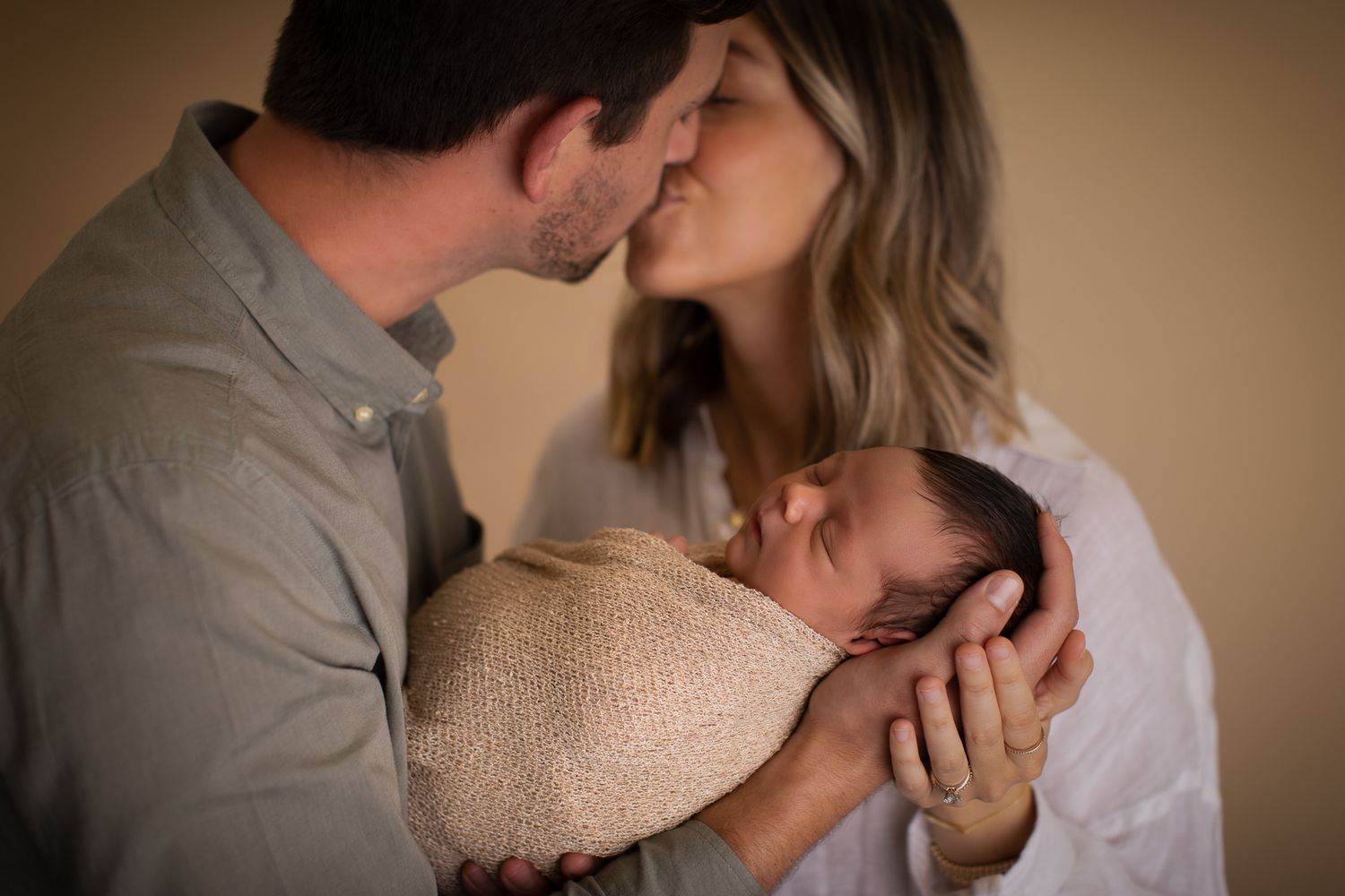 Parents sharing a kiss while cradling their swaddled newborn baby in a tender family portrait.