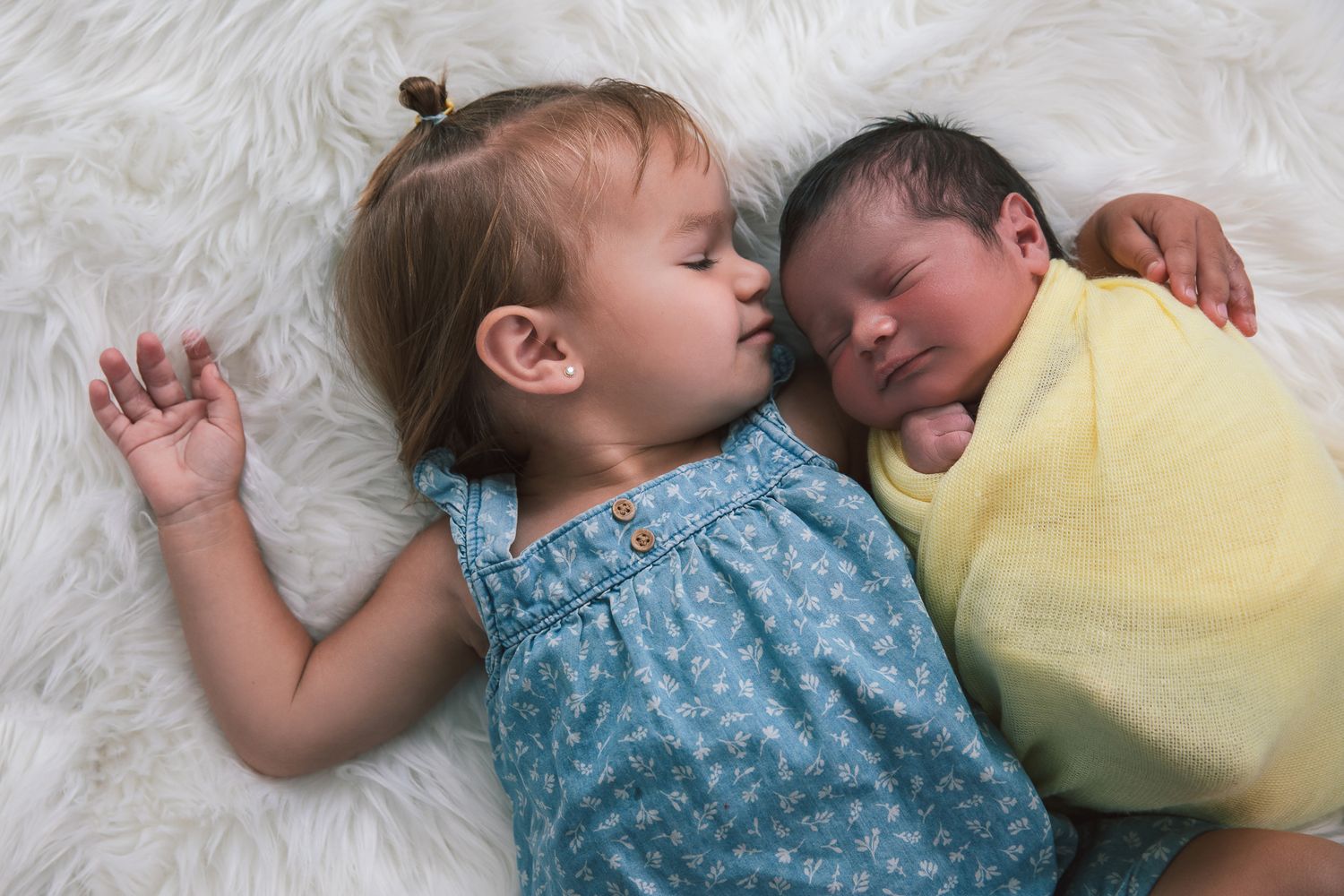 Toddler girl in blue dress sleeping beside newborn baby wrapped in yellow on a white fluffy blanket.