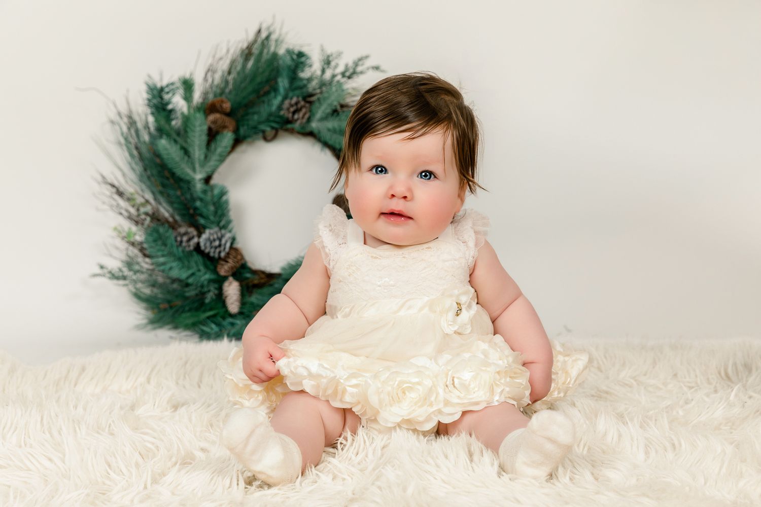 Baby girl in cream dress sits on white fur rug before holiday wreath backdrop.