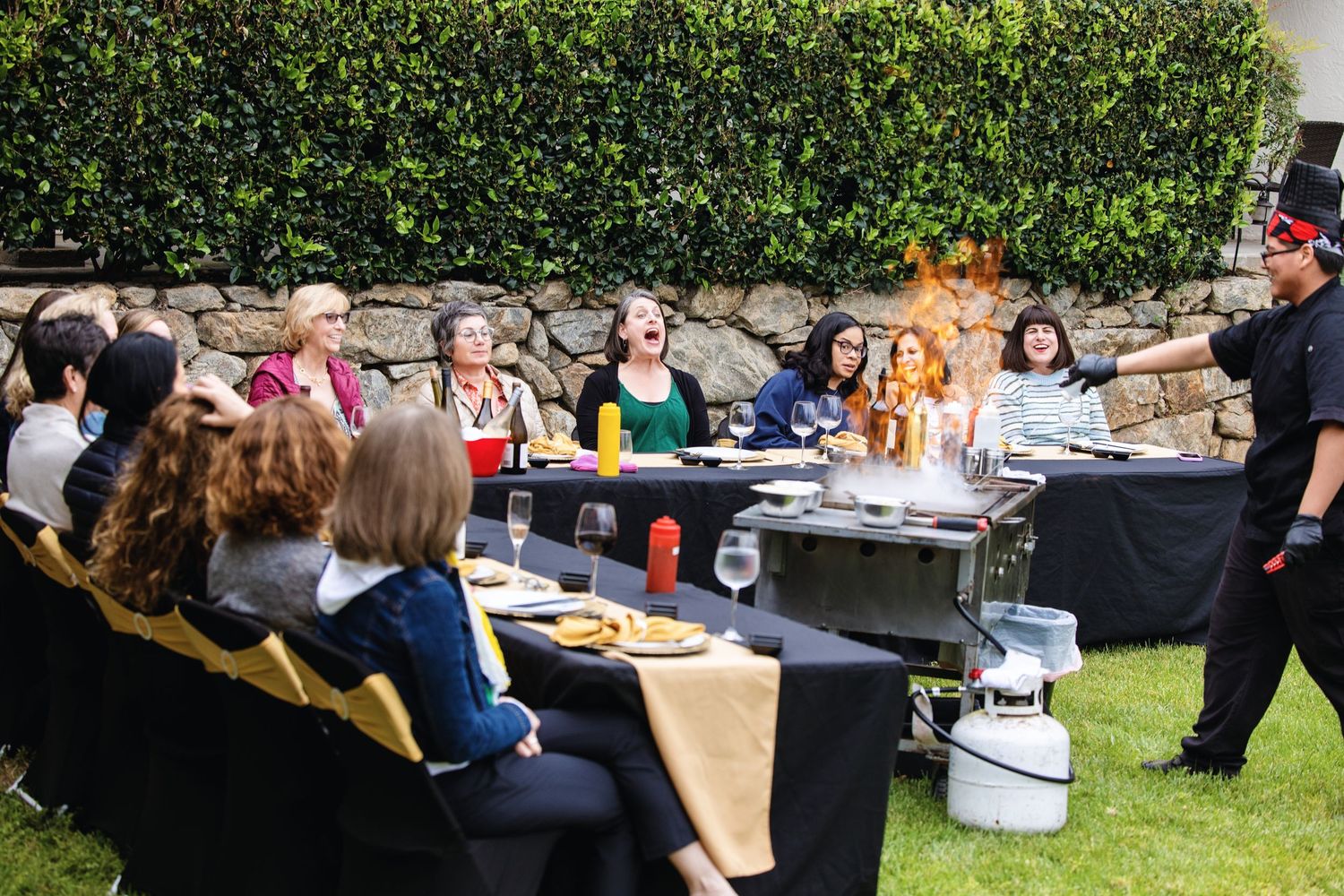Chef performs flambé tableside for excited women at outdoor dining event on green lawn.