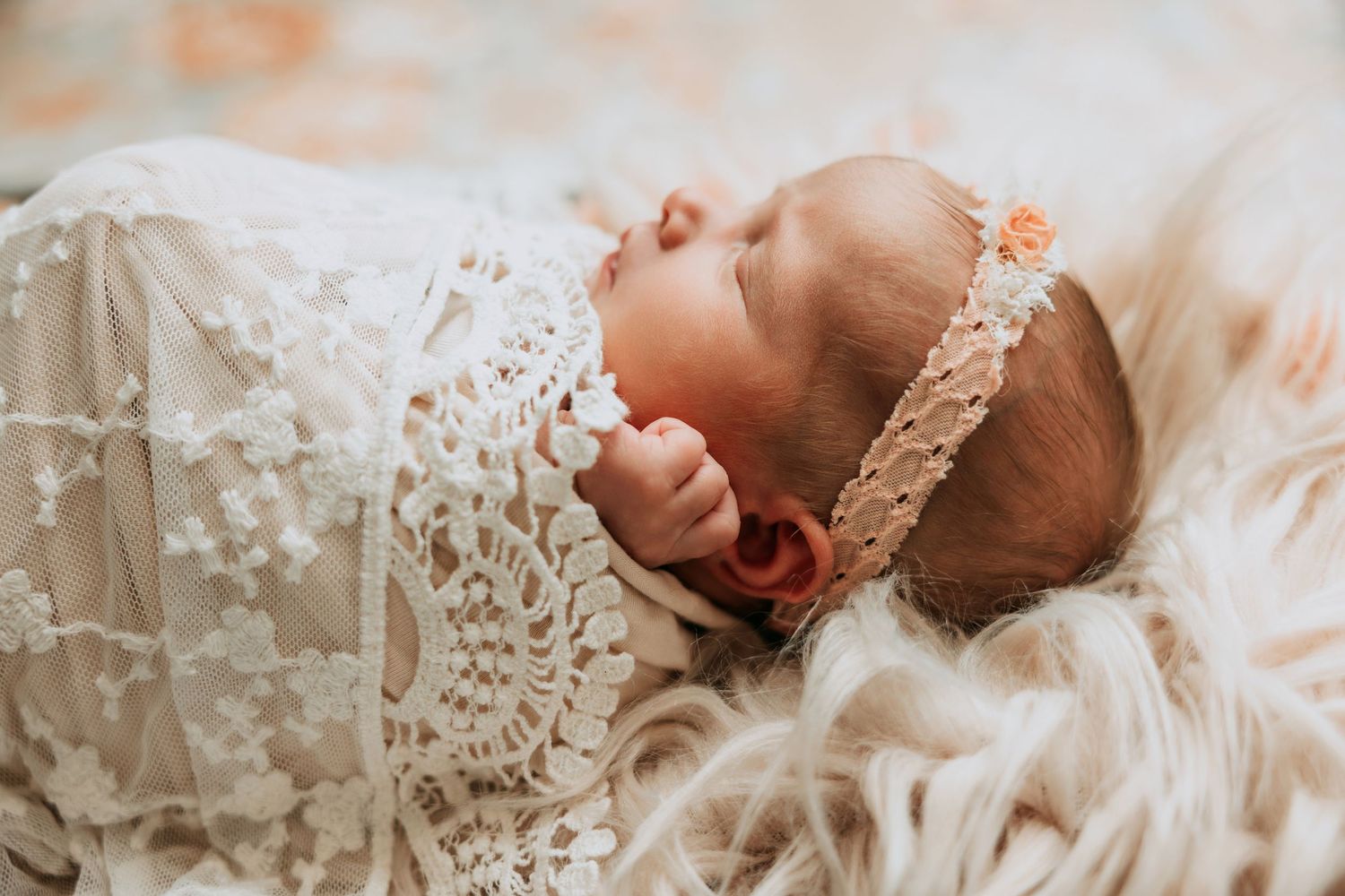 Sleeping newborn baby wrapped in white lace swaddle with floral headband on fluffy fur blanket.