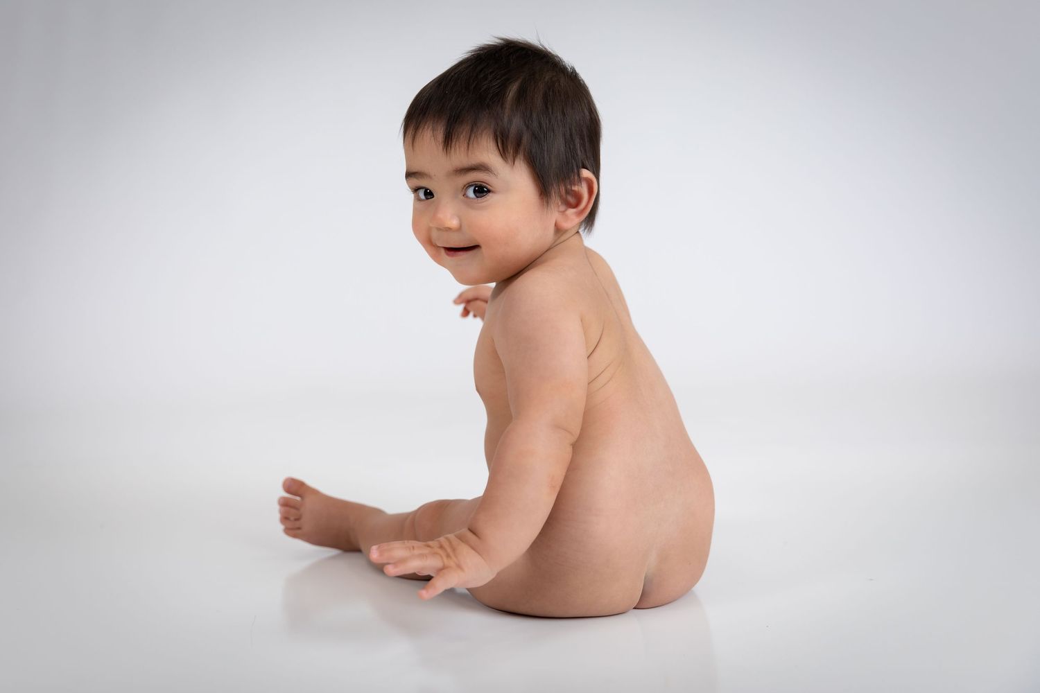 Cute baby sitting on white background, looking over shoulder with bright eyes and happy expression.