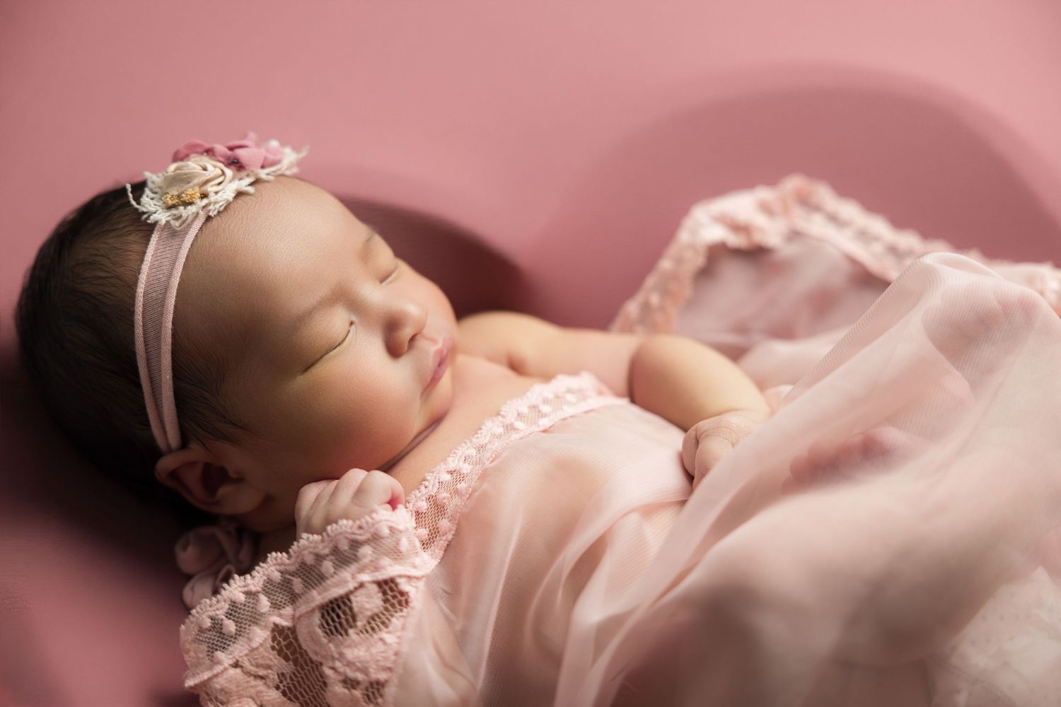 Sleeping newborn baby girl in pink lace outfit and floral headband on pink background.