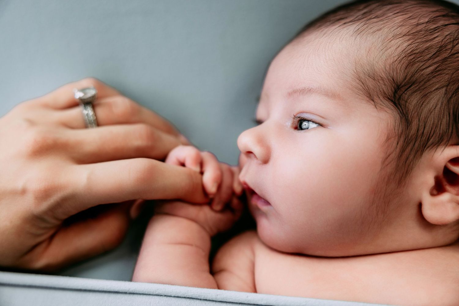 Newborn baby grasping mother's finger in a white crib, with mother wearing a diamond ring.